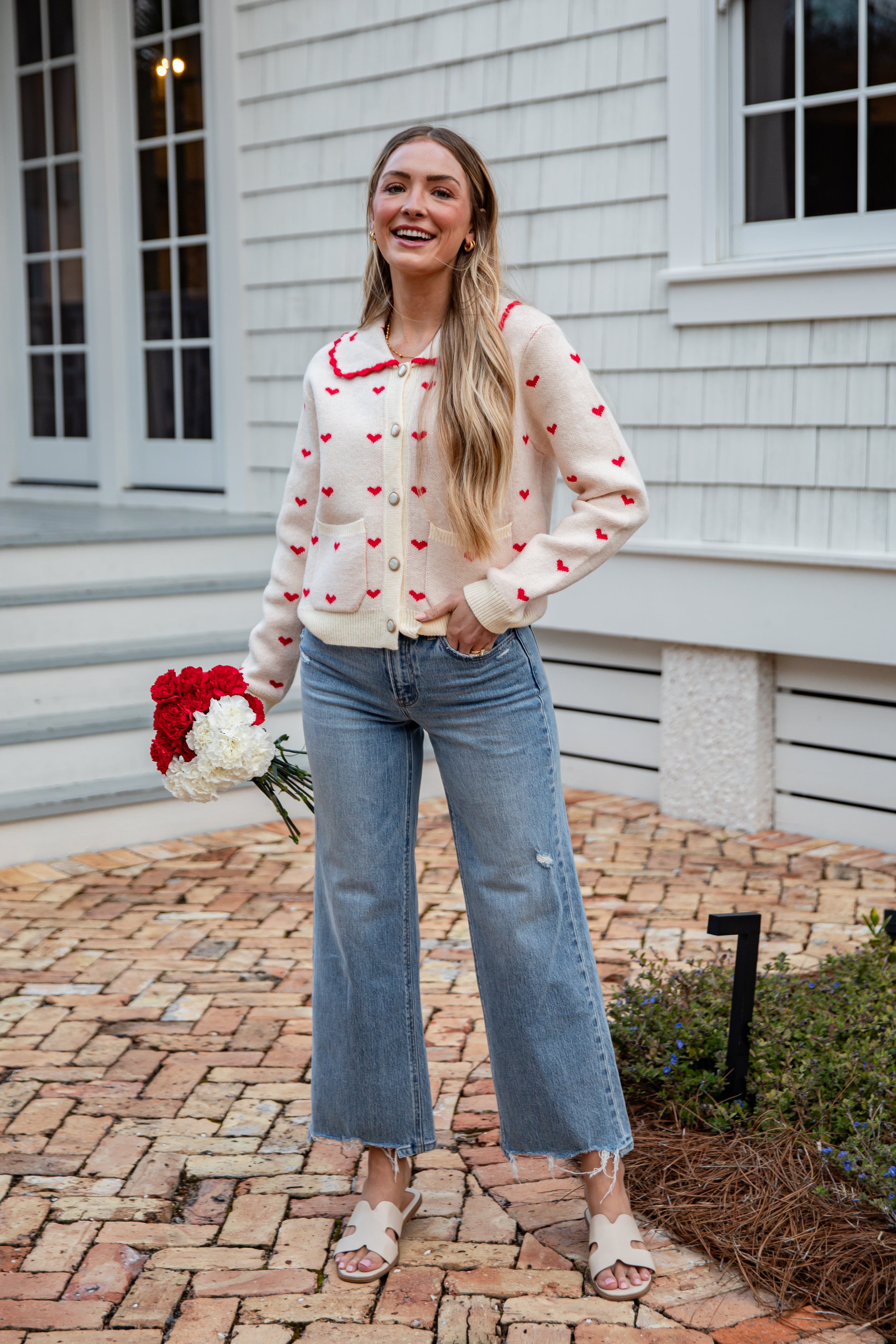 Woman wearing a heart-patterned cardigan and jeans, standing on a brick path in front of a house.