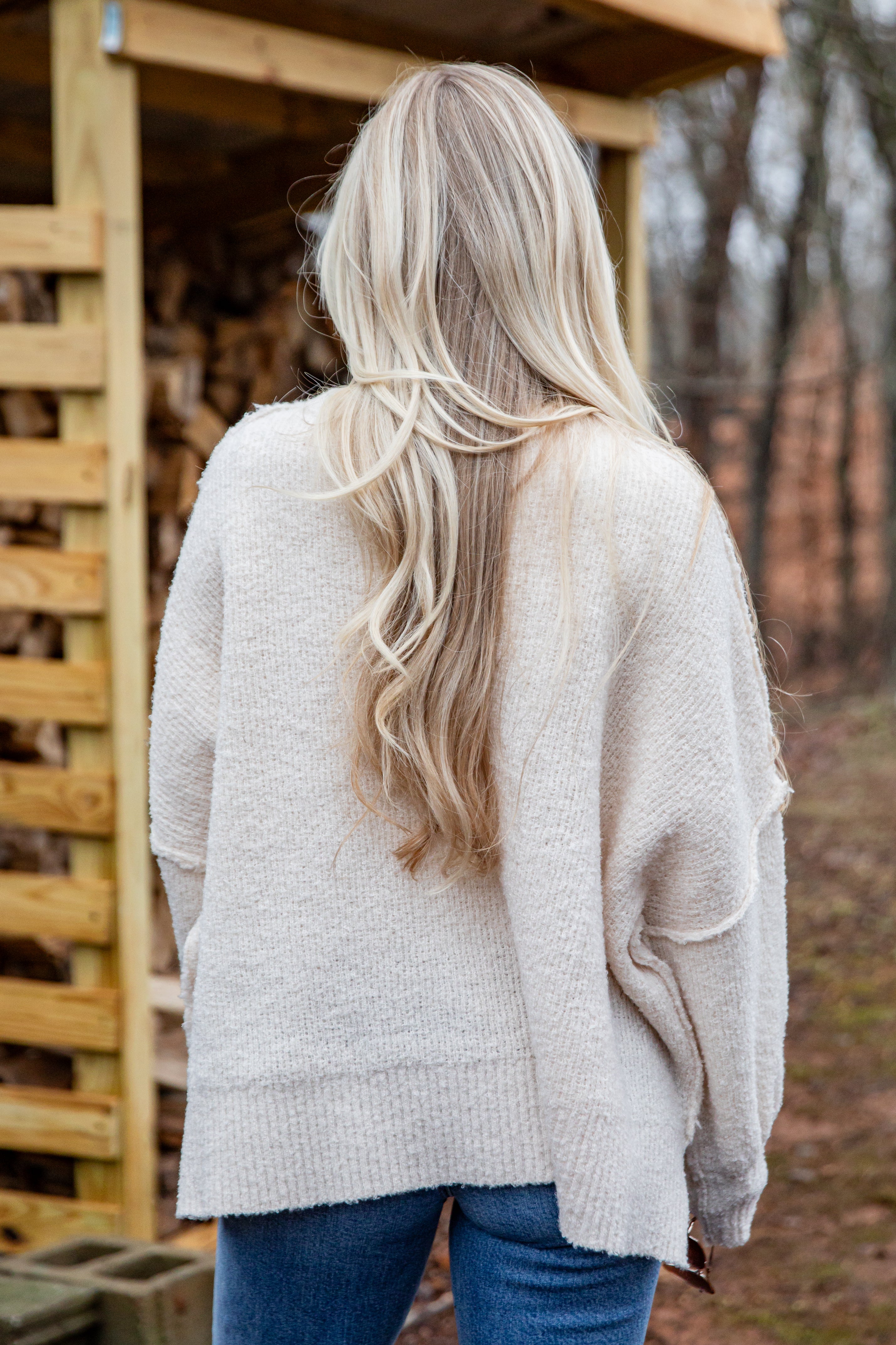 Person wearing a light gray sweater with long blonde hair, standing in a rustic outdoor setting.
