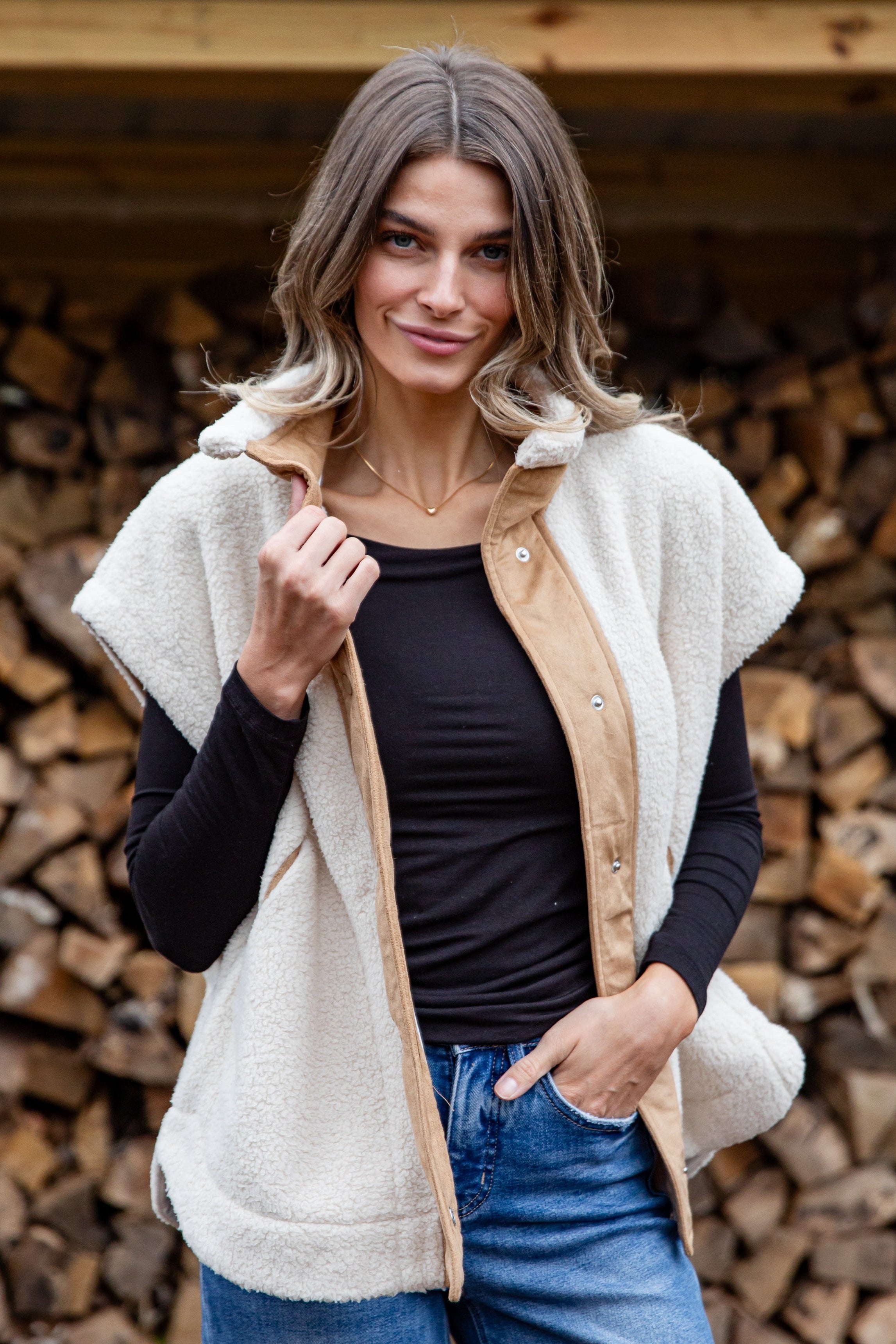 Woman wearing a beige vest over a black top and blue jeans, standing in front of stacked firewood.