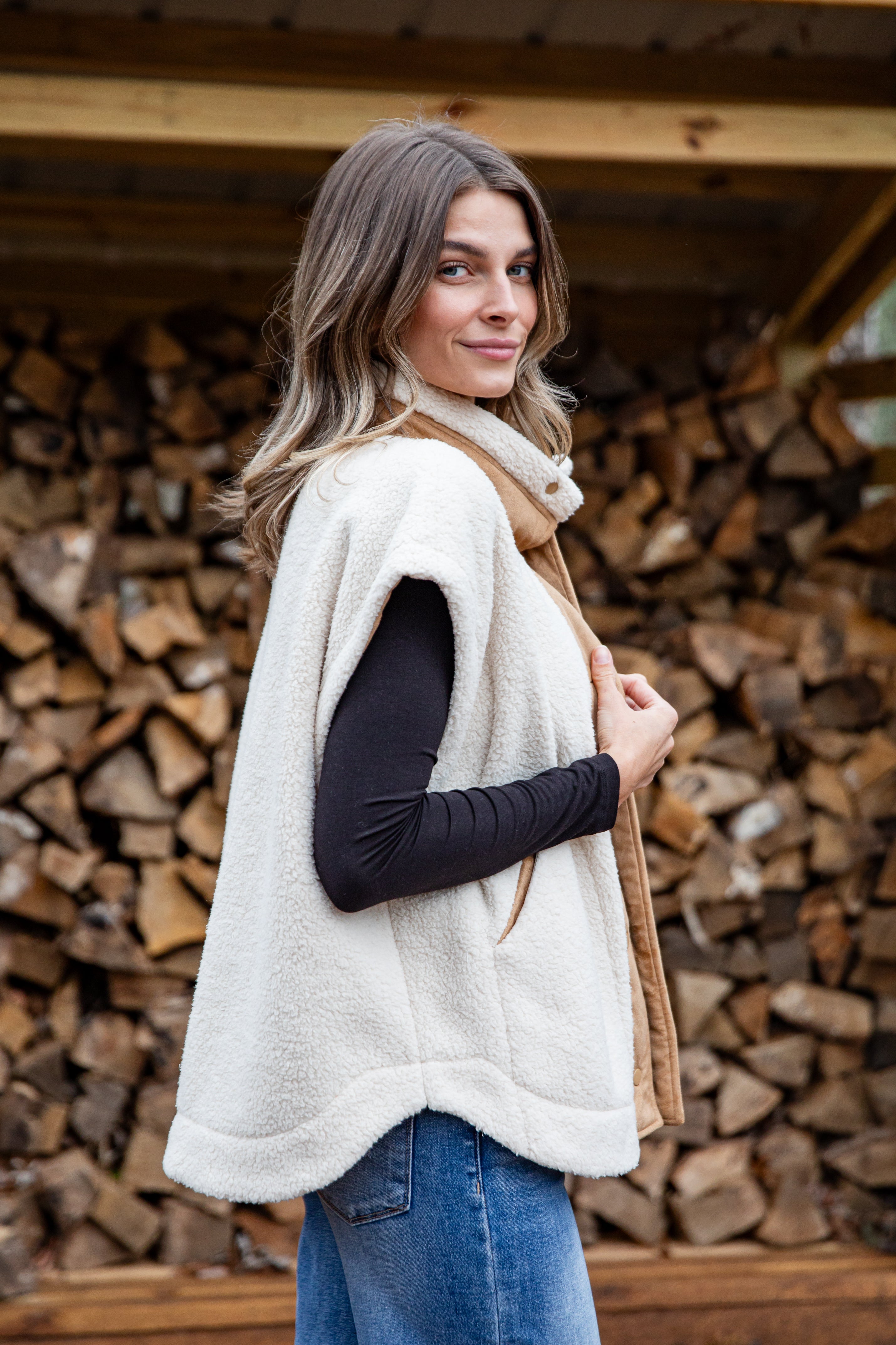 Woman wearing a white vest over a black long-sleeve shirt with a beige scarf, standing in front of stacked firewood.
