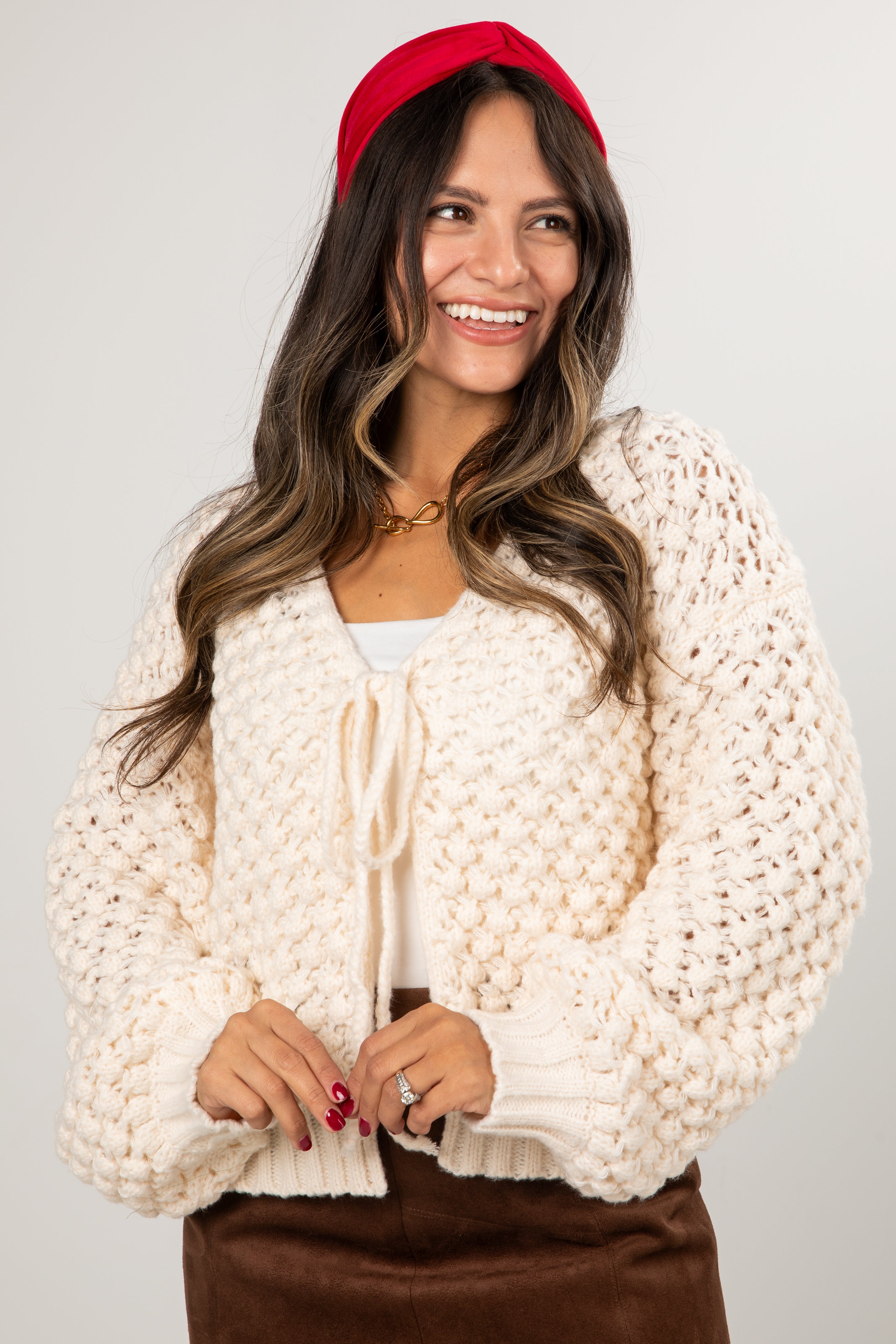 Woman wearing a cream chunky knit cardigan over a white top with a red headband on a plain background