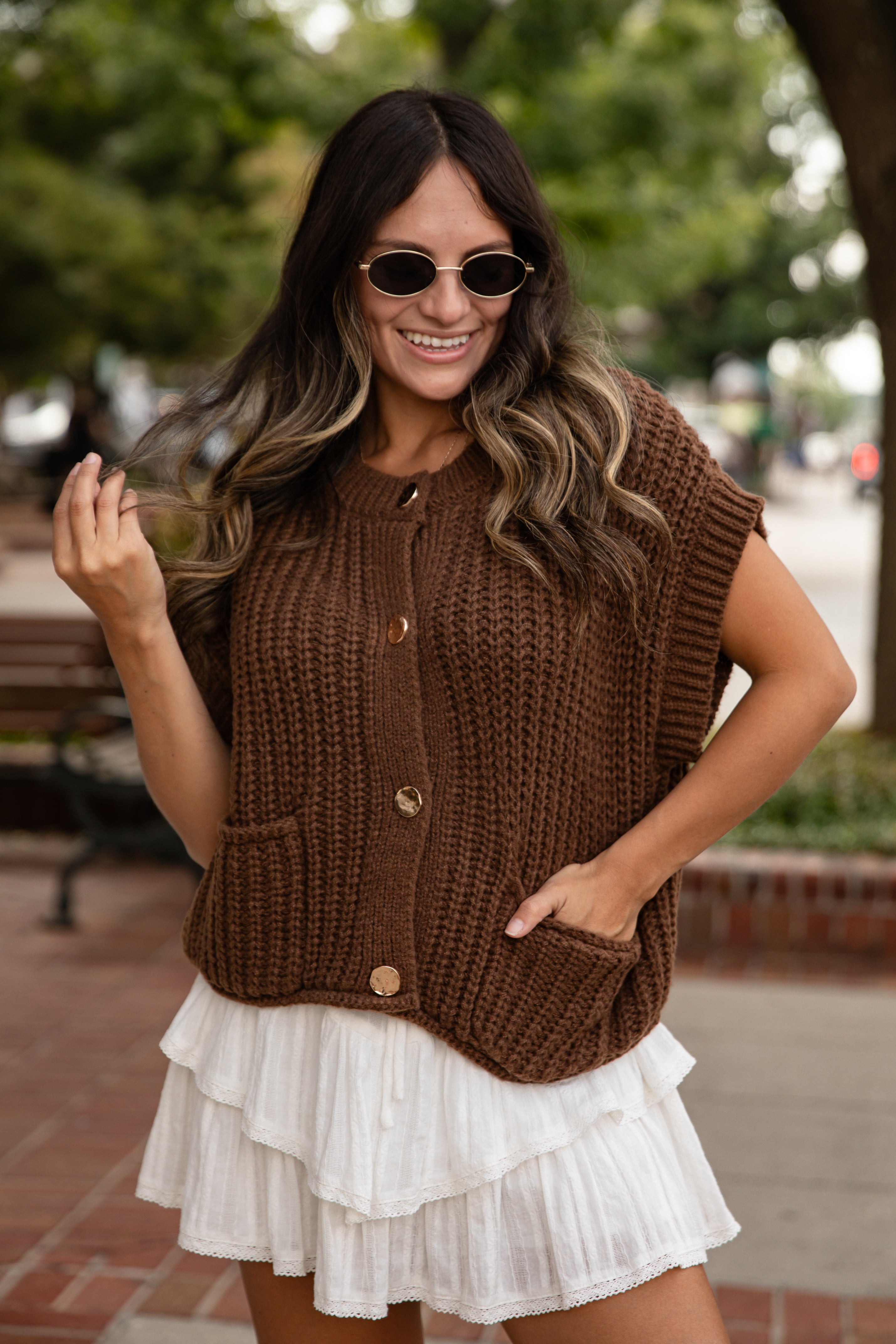 Woman wearing a brown knitted vest over a white dress outdoors.