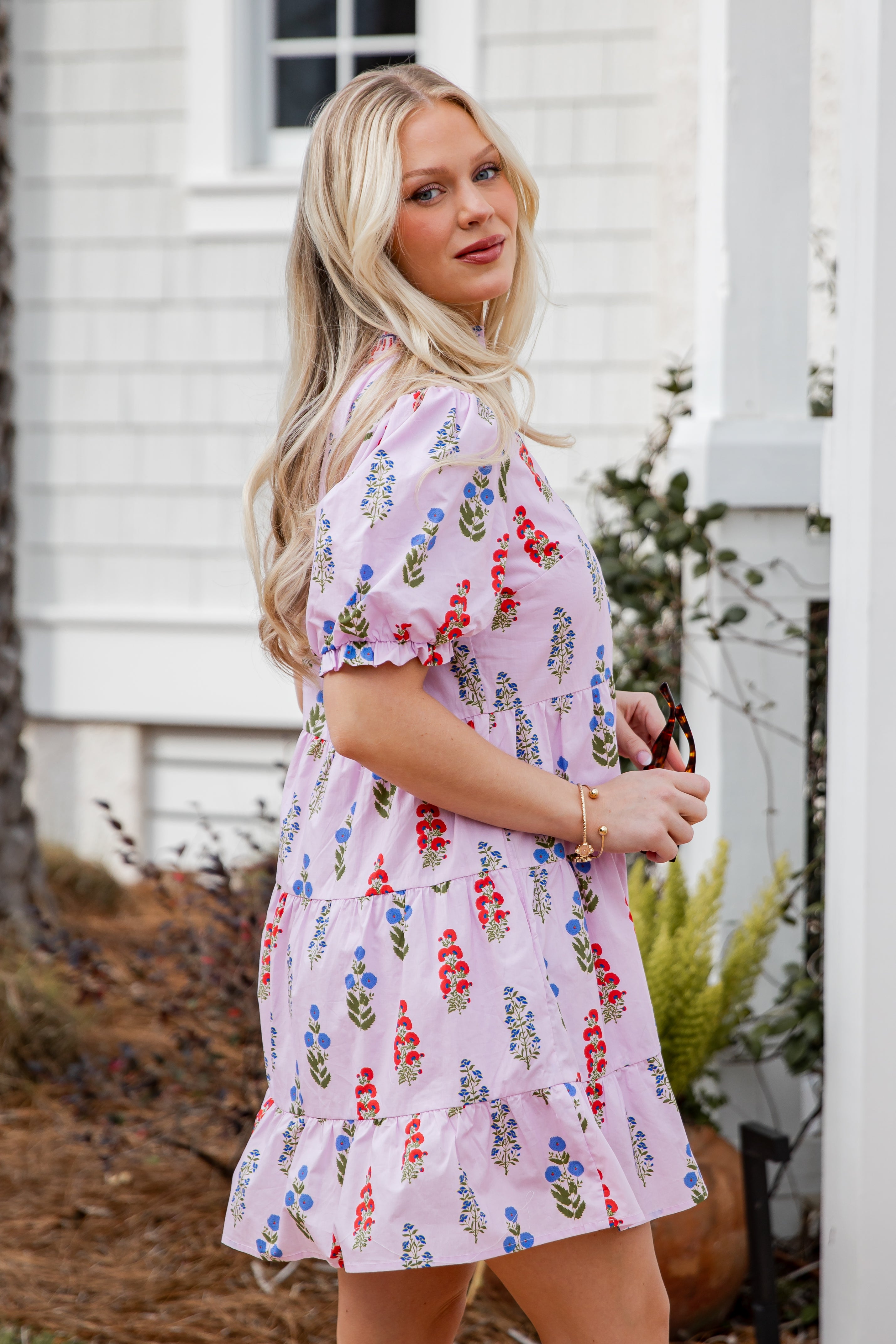 Woman wearing a floral dress standing outdoors with a white building and plants in the background