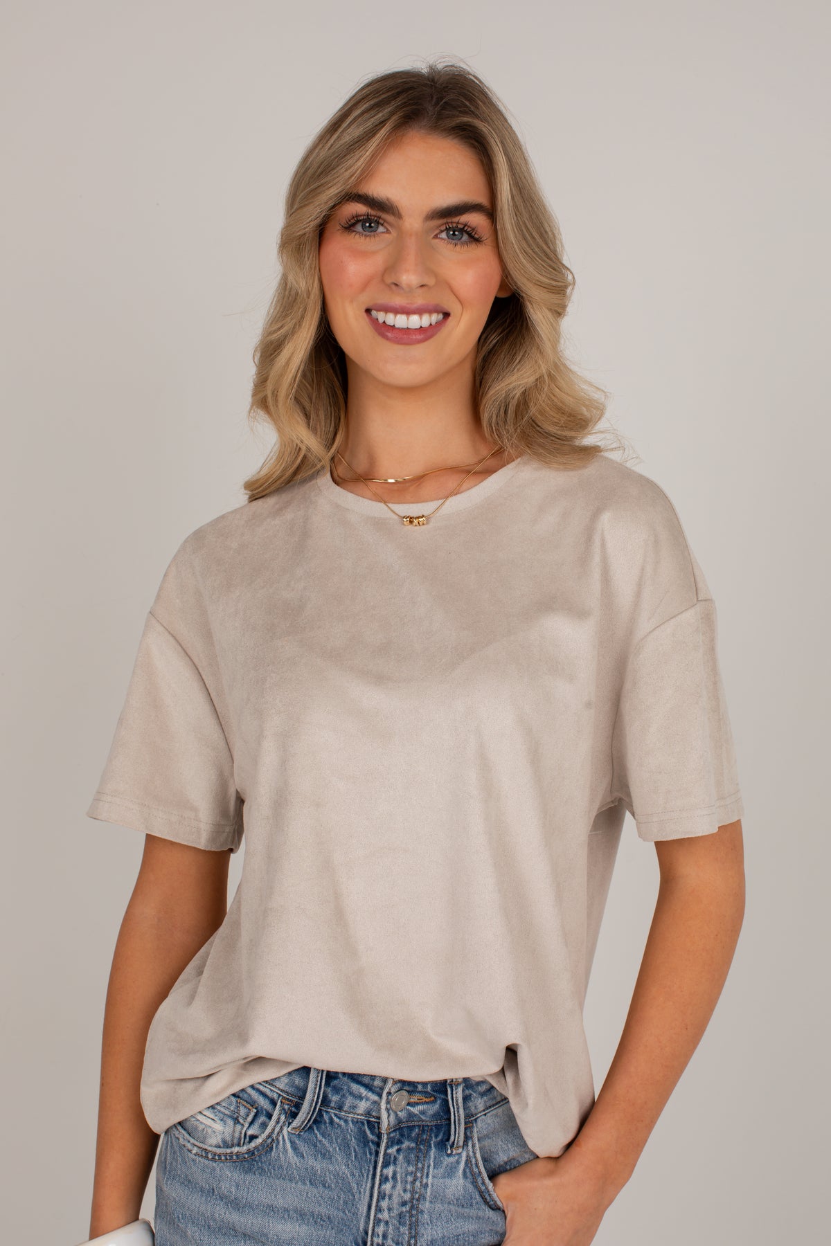 A smiling blonde woman wearing a light beige short-sleeve V-neck t-shirt and blue jeans, standing against a neutral light gray background.