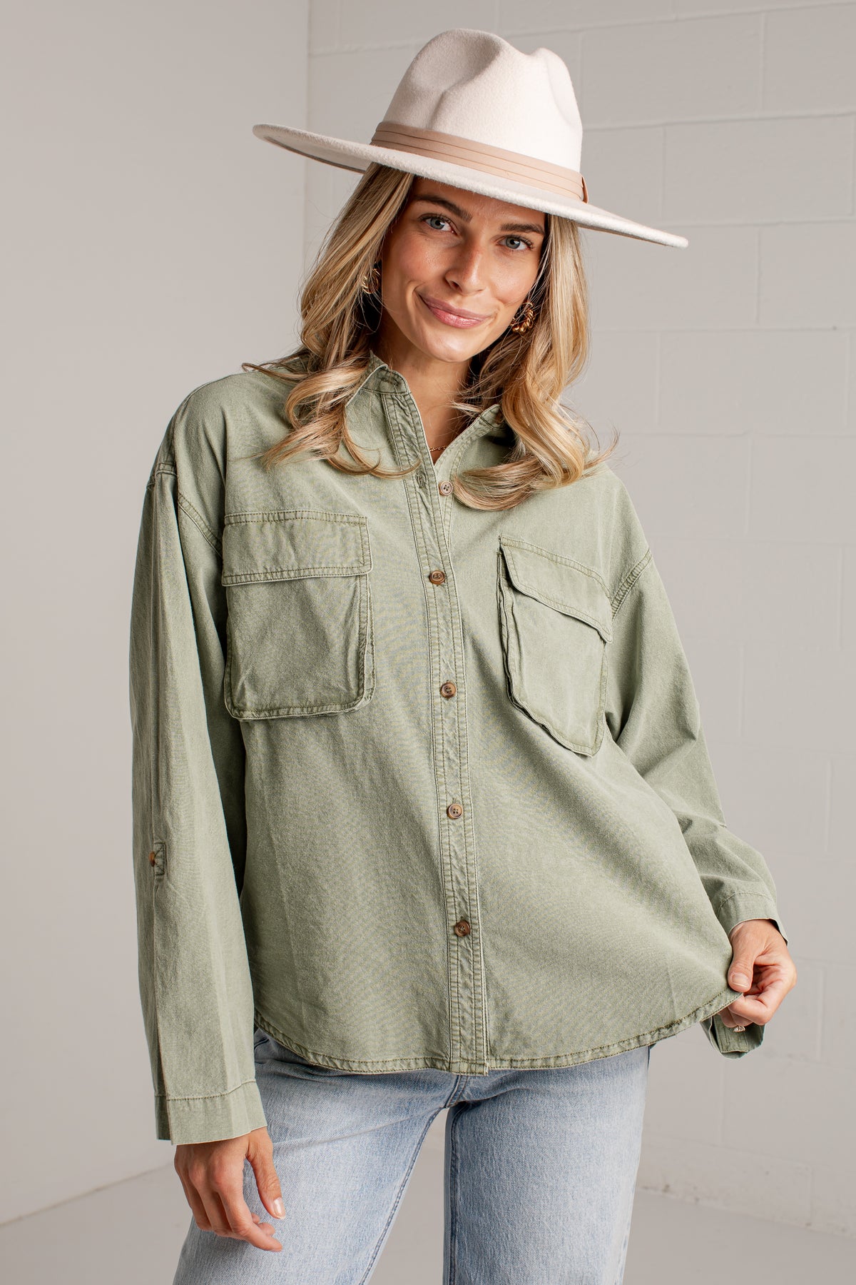 A woman wearing an oversized sage green button-up shirt with two chest pockets, paired with light-wash jeans and a wide-brimmed cream hat, standing against a light gray brick wall.