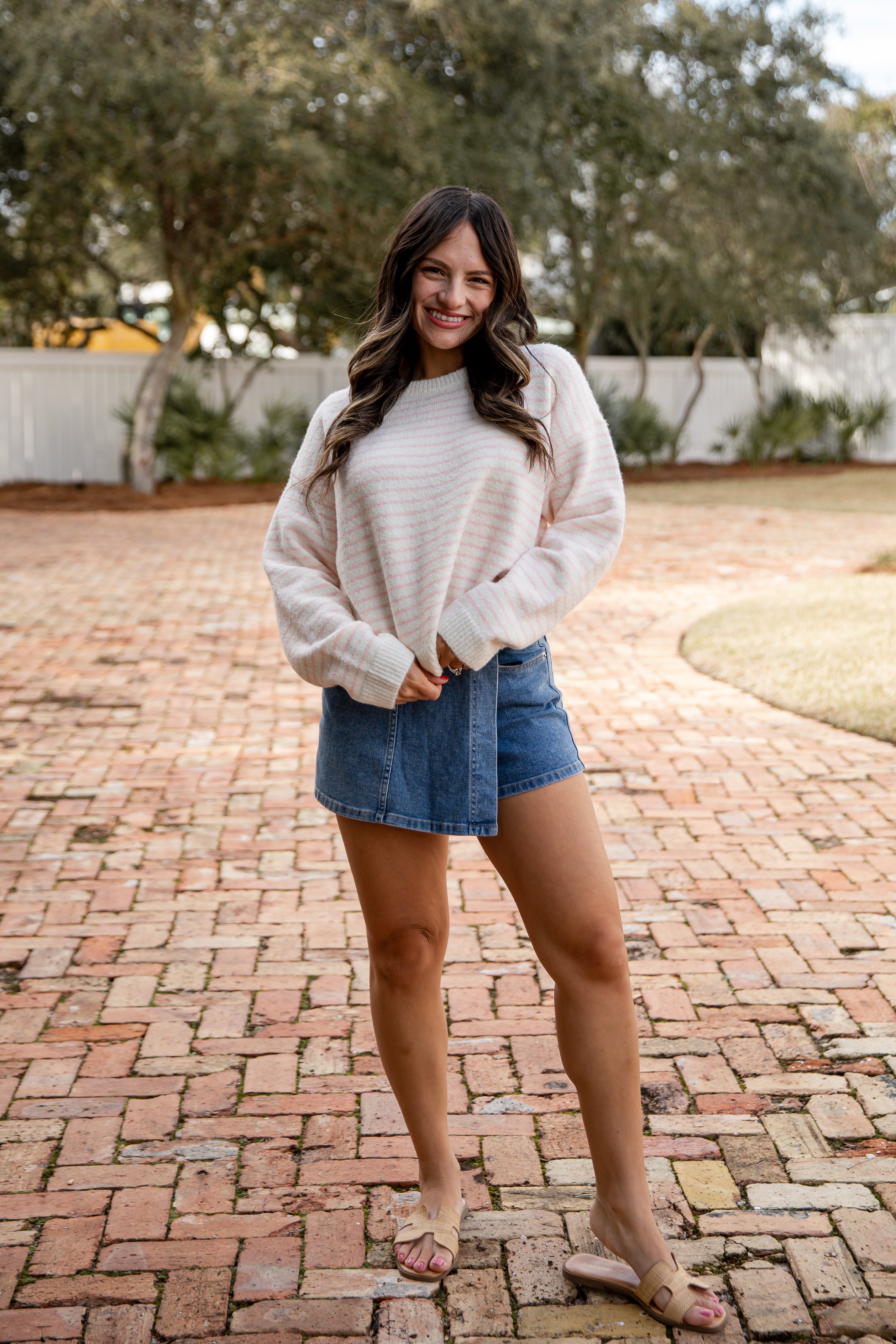 Woman wearing a white sweater and denim shorts standing on a brick patio with trees in the background