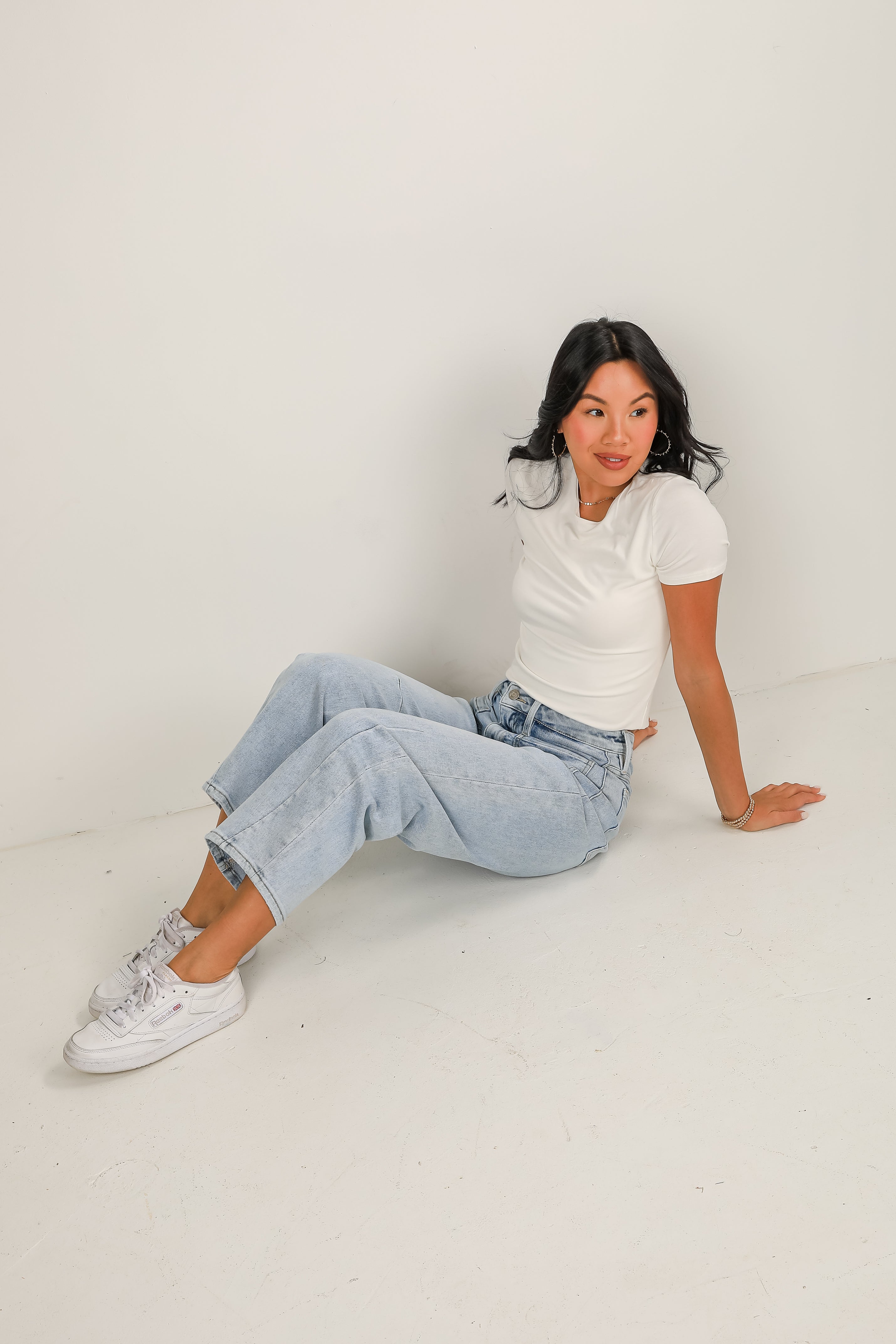 Woman sitting on a white floor wearing a white t-shirt, light blue jeans, and white sneakers.