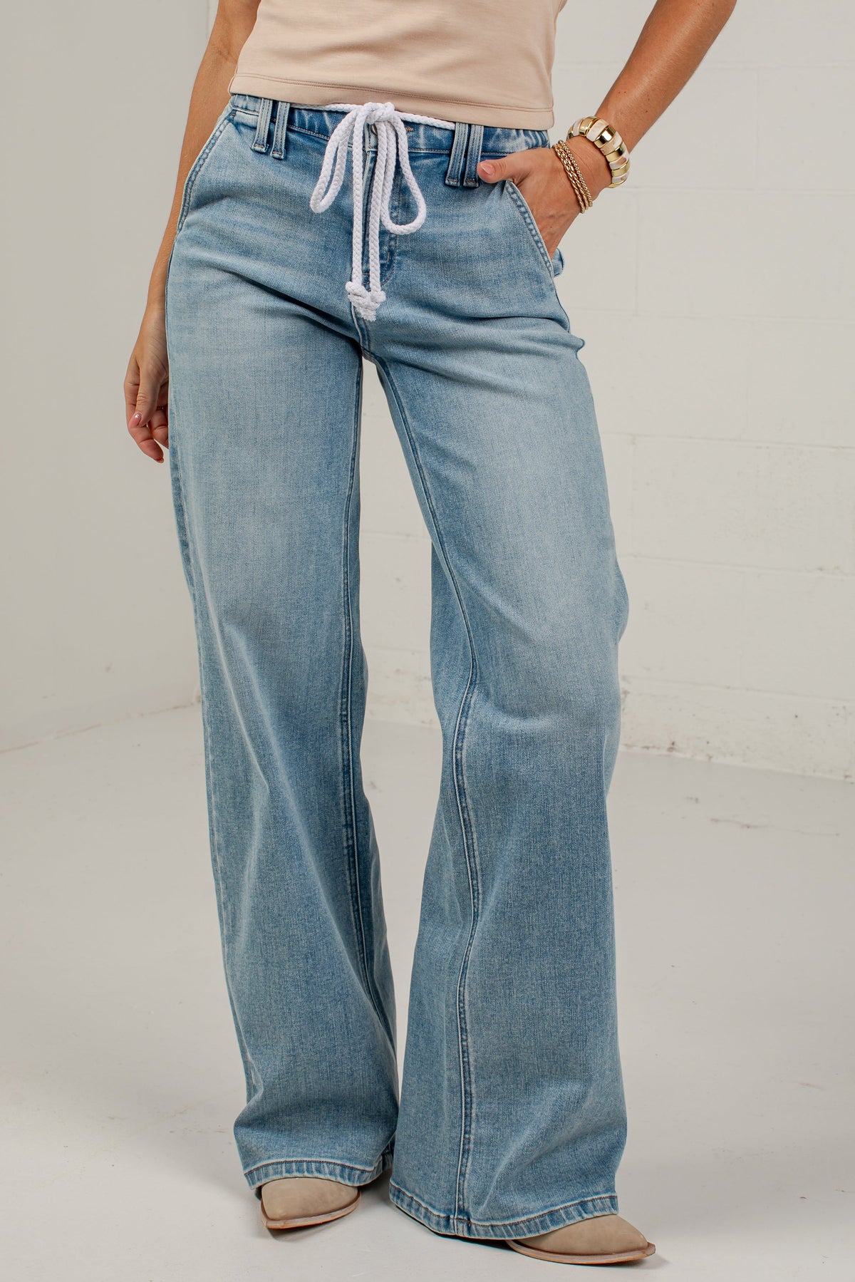 A model wearing light-wash blue wide-leg jeans with a white drawstring waist, paired with a beige top and beige shoes, standing against a white studio background.