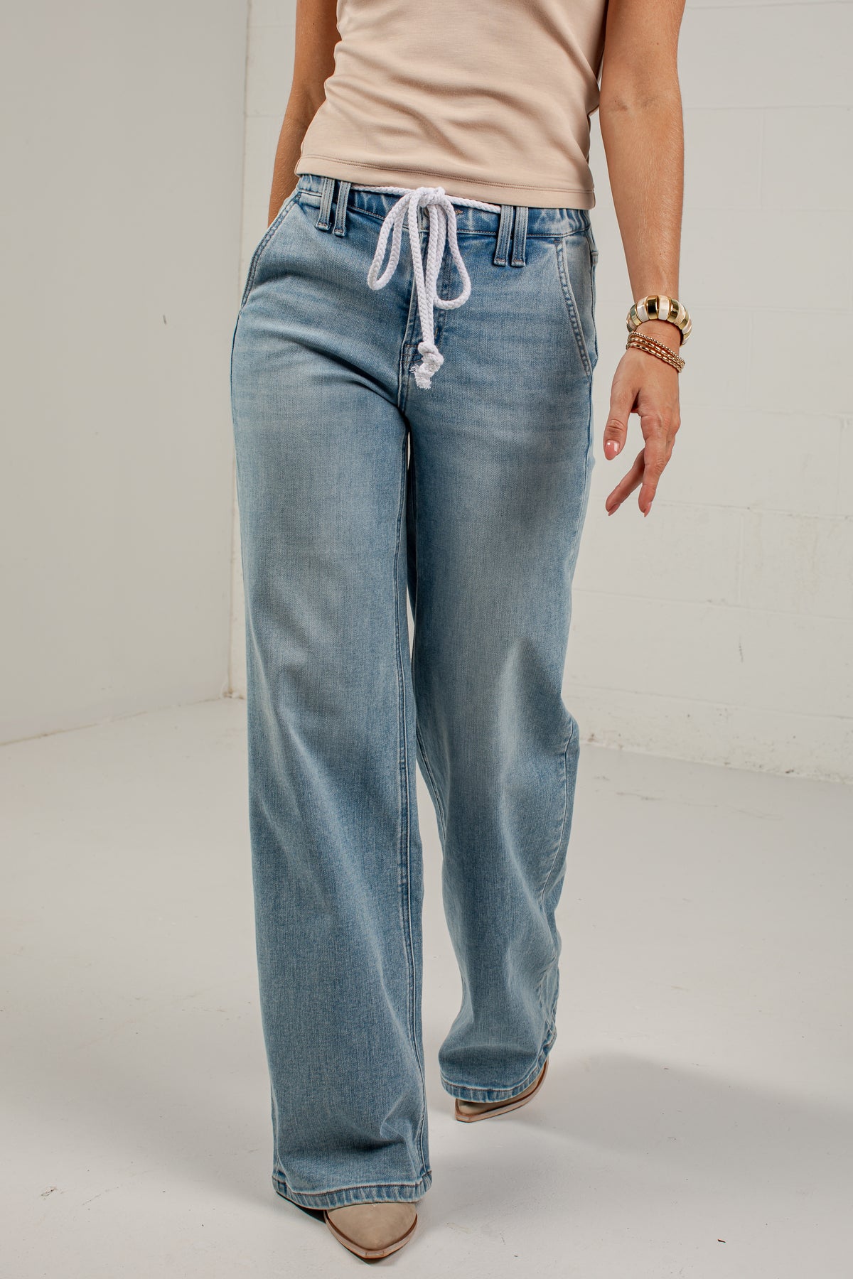 A person wearing light-wash blue wide-leg jeans with a white drawstring waist, paired with a beige top and beige shoes, standing against a plain white studio background.