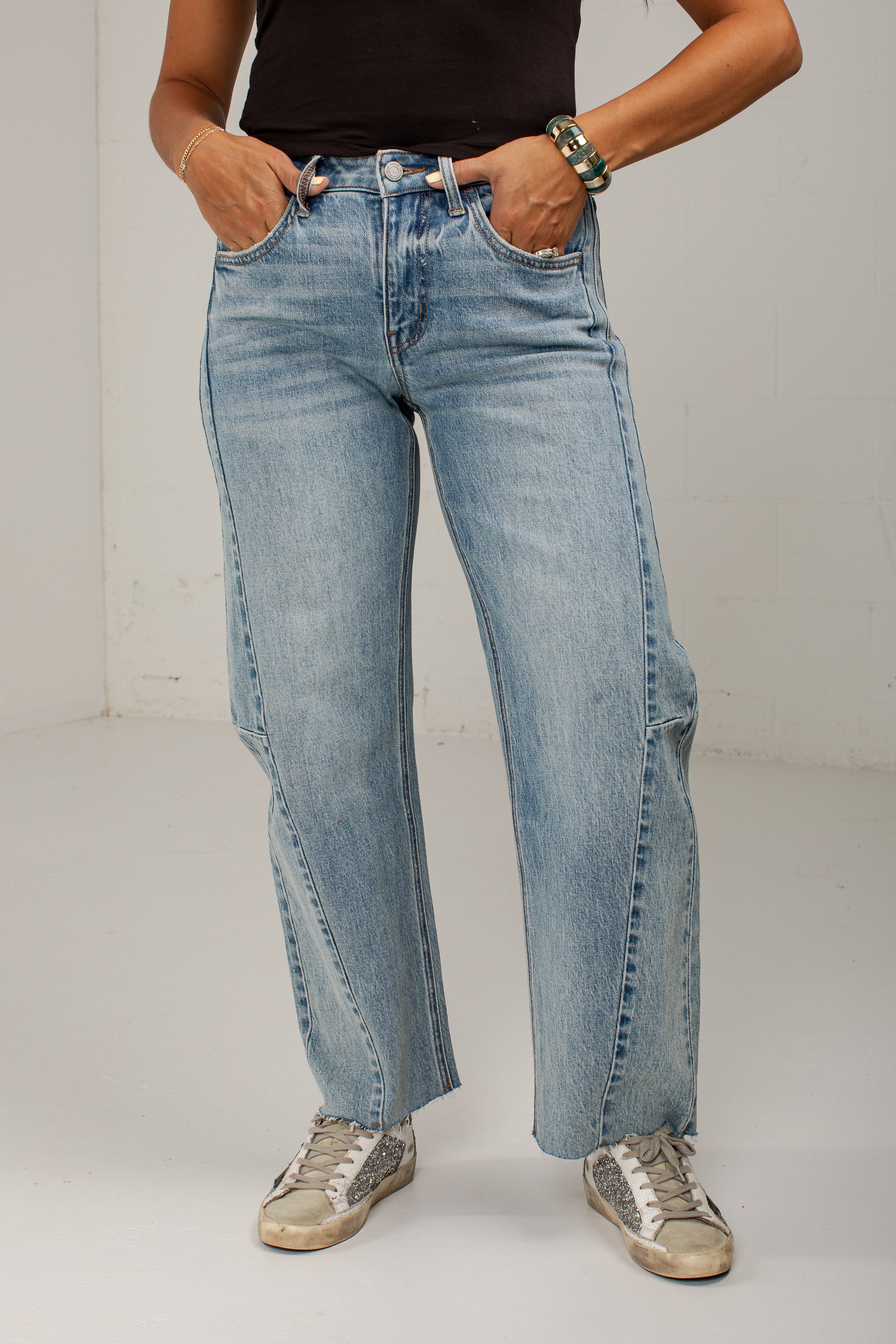 A person wearing light-wash blue straight-leg jeans with side cargo pockets, paired with a black top and distressed sneakers, standing against a plain light gray wall.