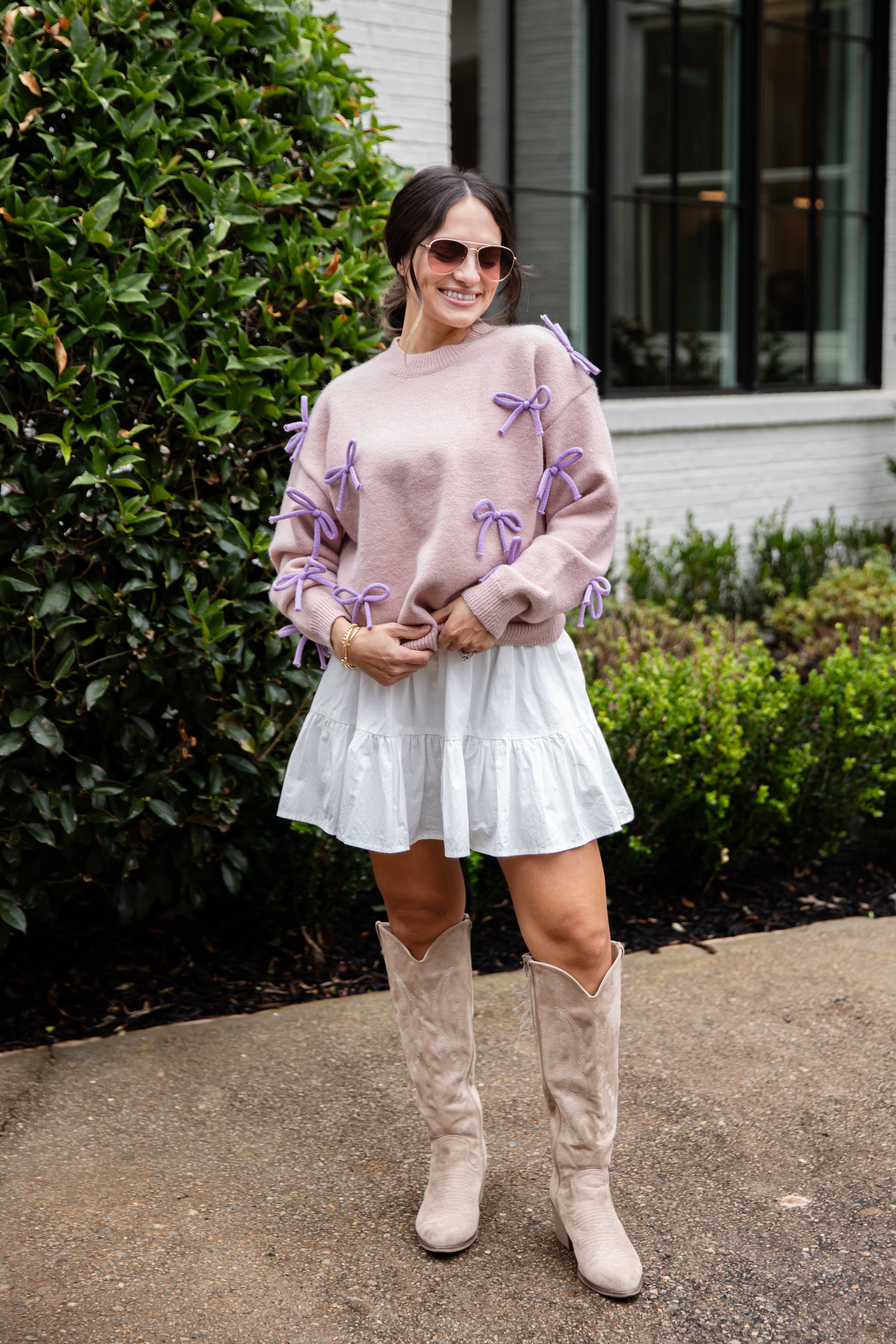 Woman wearing a pink sweater with purple bows, white skirt, and beige knee-high boots standing outdoors.