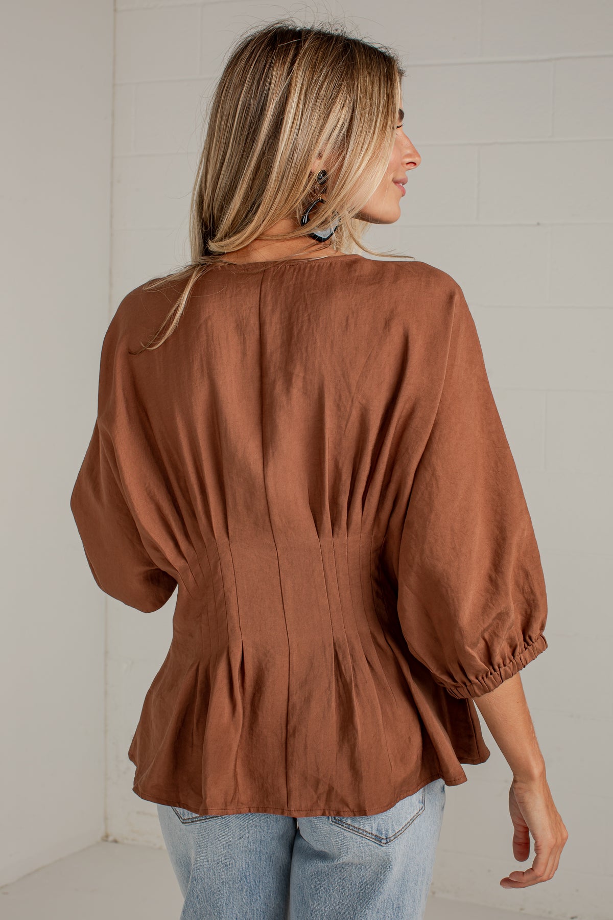 A woman viewed from the back wearing a rust-brown long-sleeve blouse with gathered puff sleeves and a peplum hem, paired with light blue jeans against a white paneled wall.