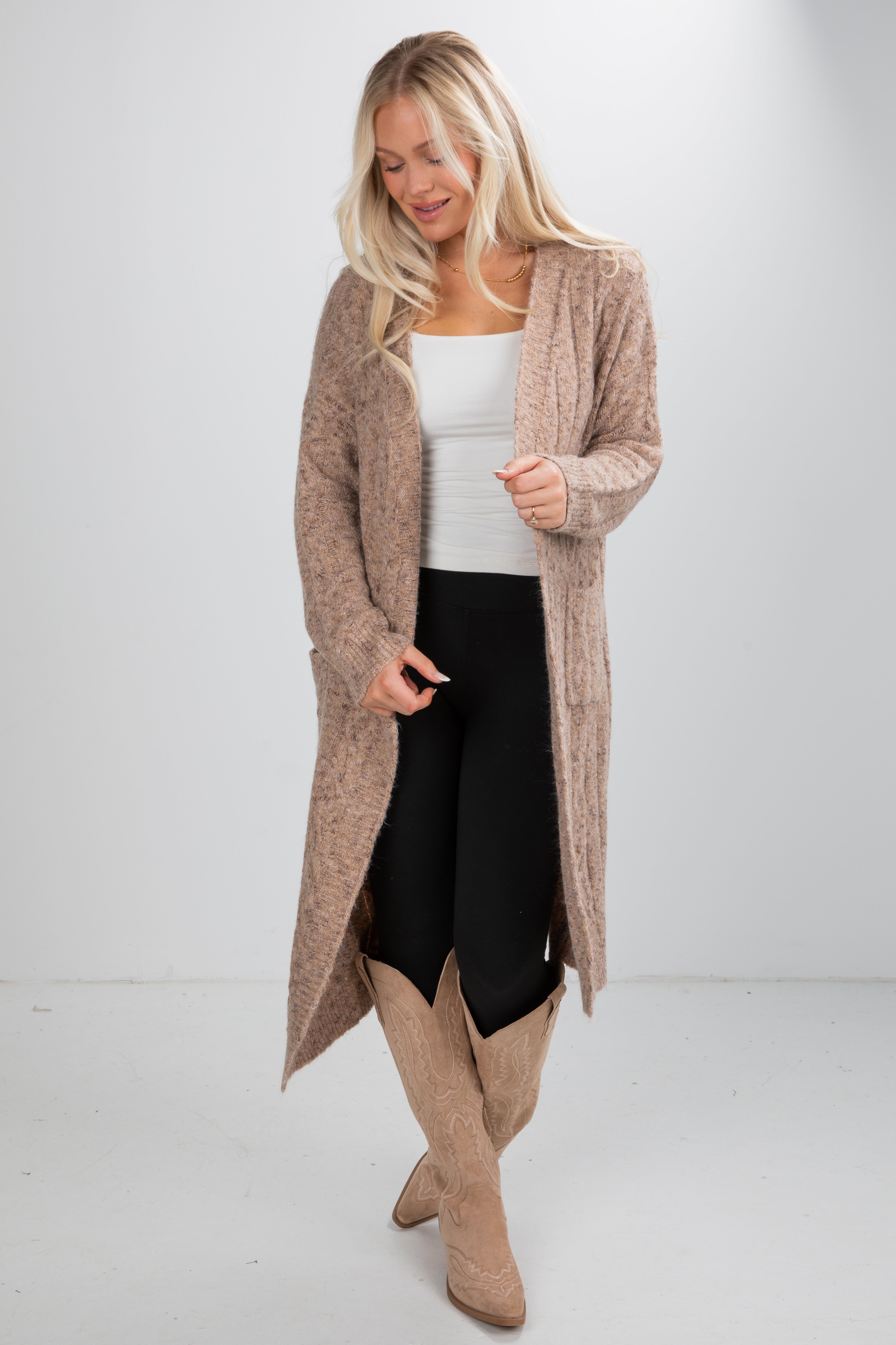 Woman wearing a long beige cardigan, white top, black pants, and tan boots on a white background