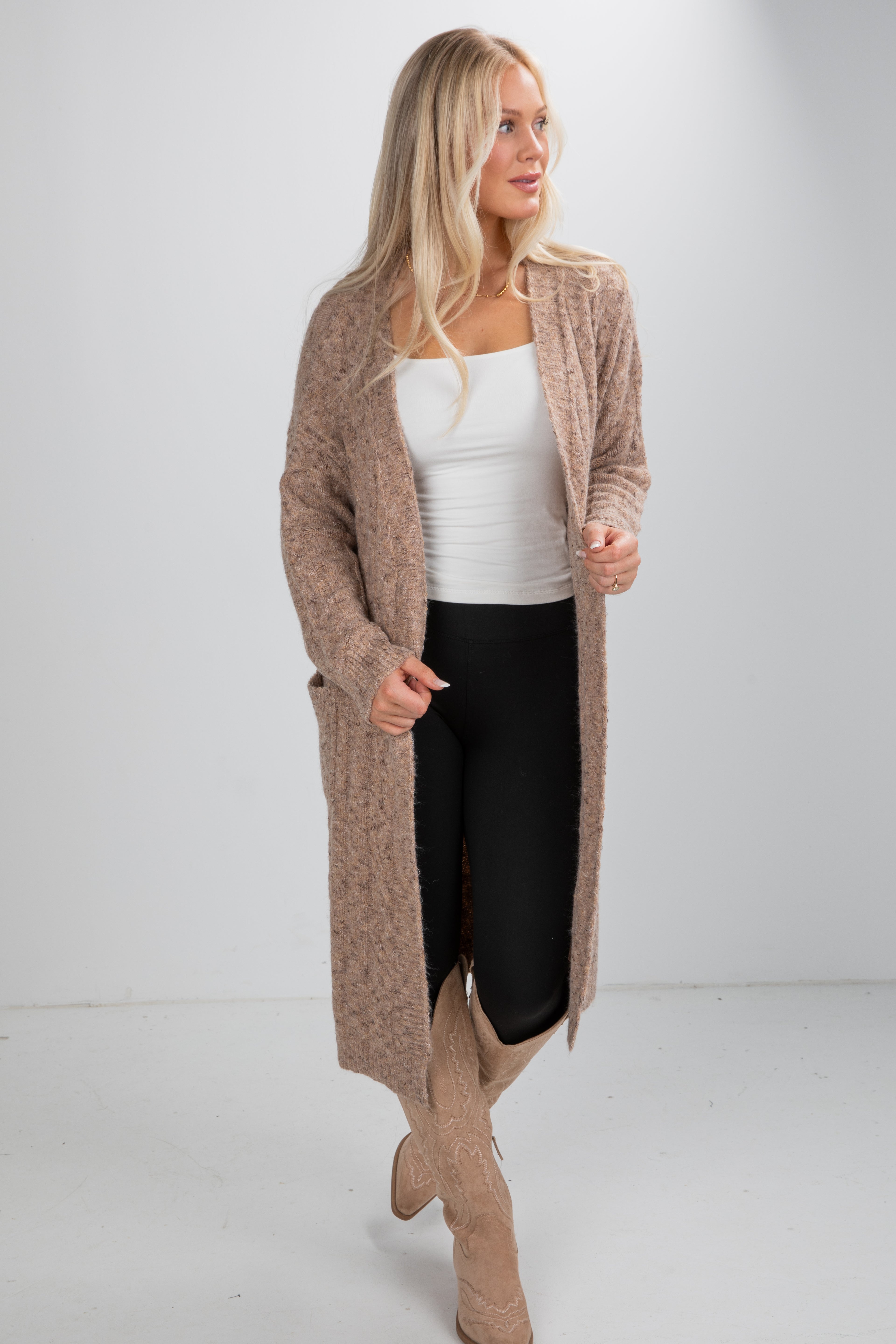 Woman wearing a long beige cardigan over a white top and black pants on a plain background