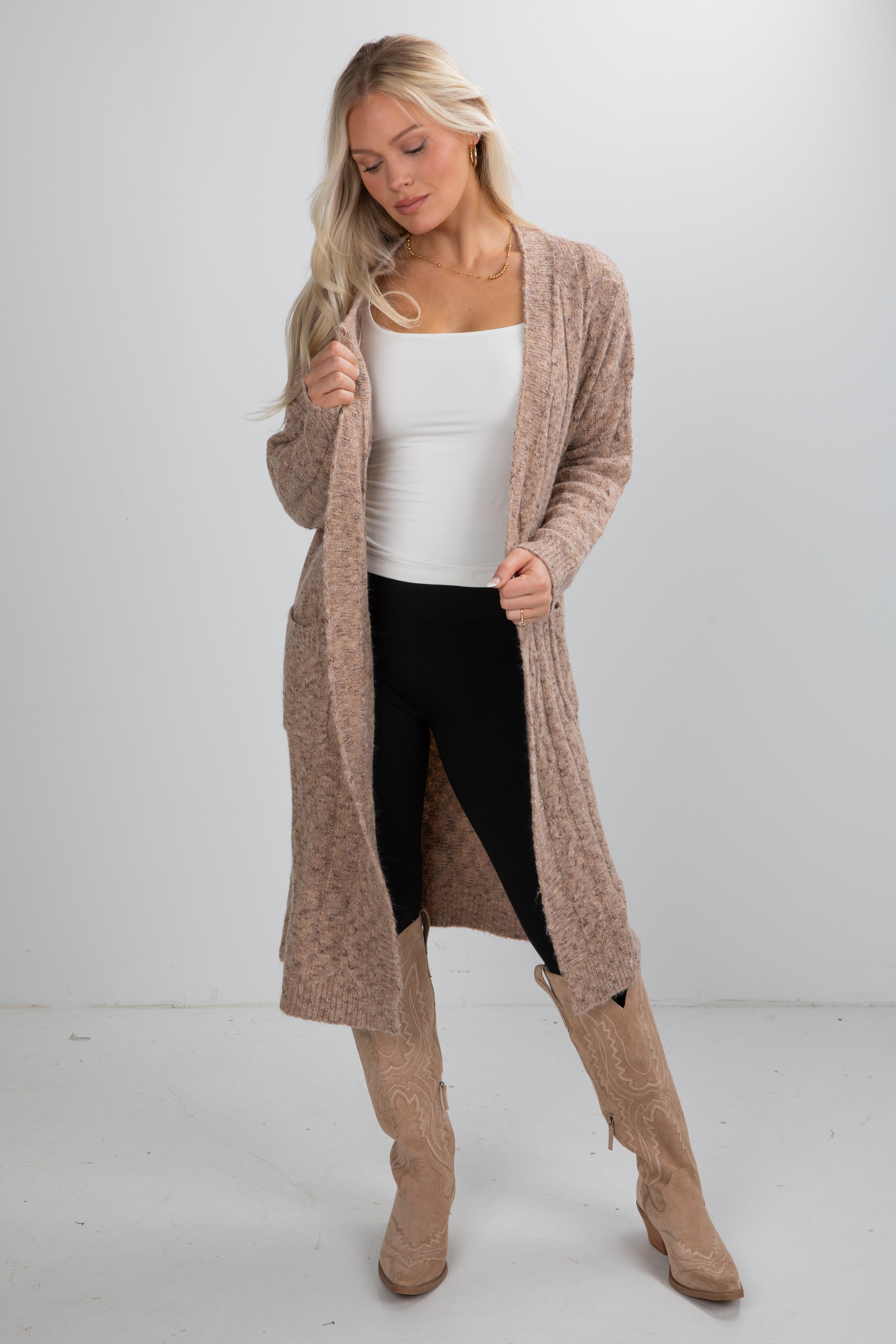 Woman wearing a long beige cardigan, white top, black leggings, and tan knee-high boots on a white background
