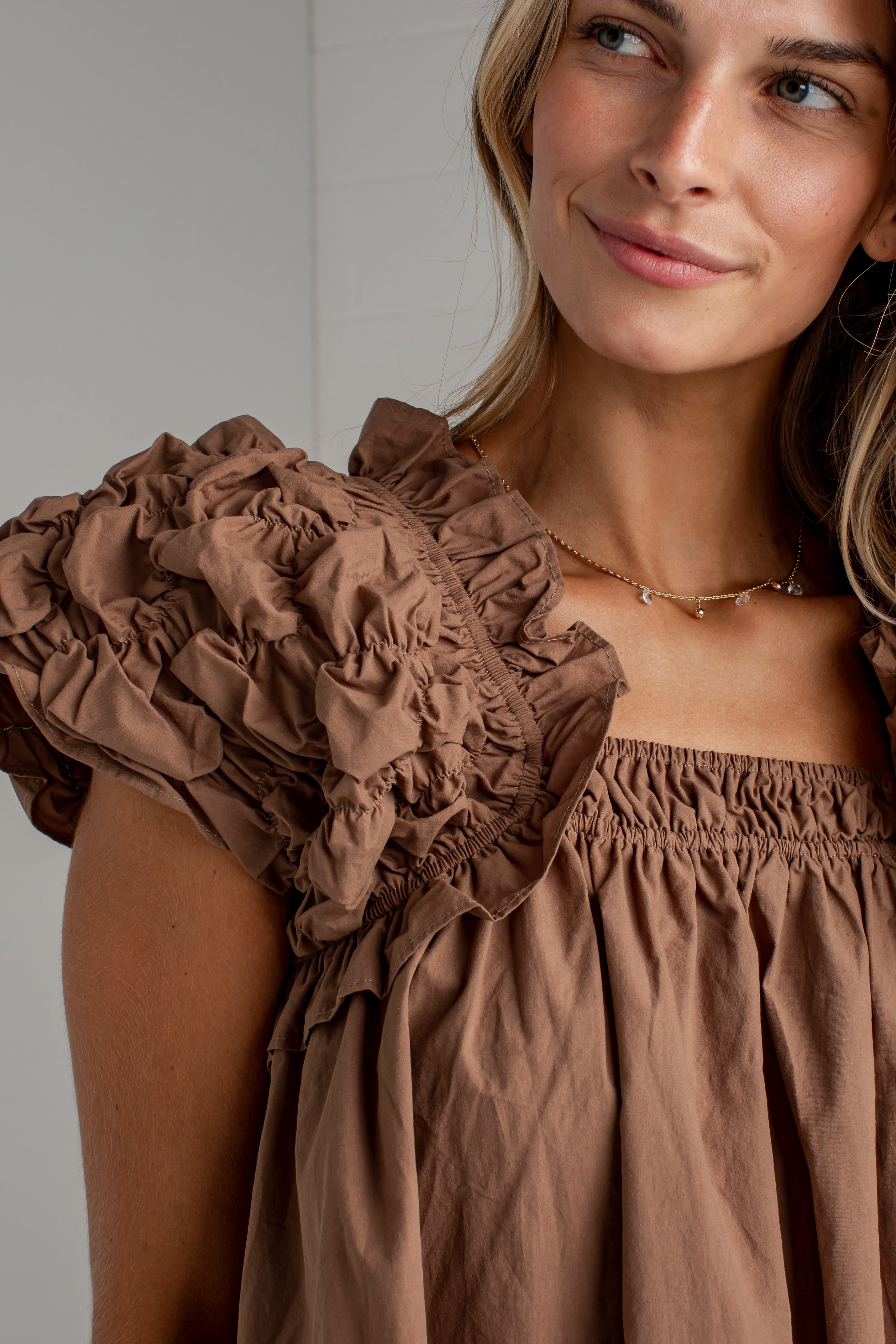 A close-up of a woman wearing a brown off-the-shoulder top with dramatic ruffled puff sleeves and a gathered neckline.