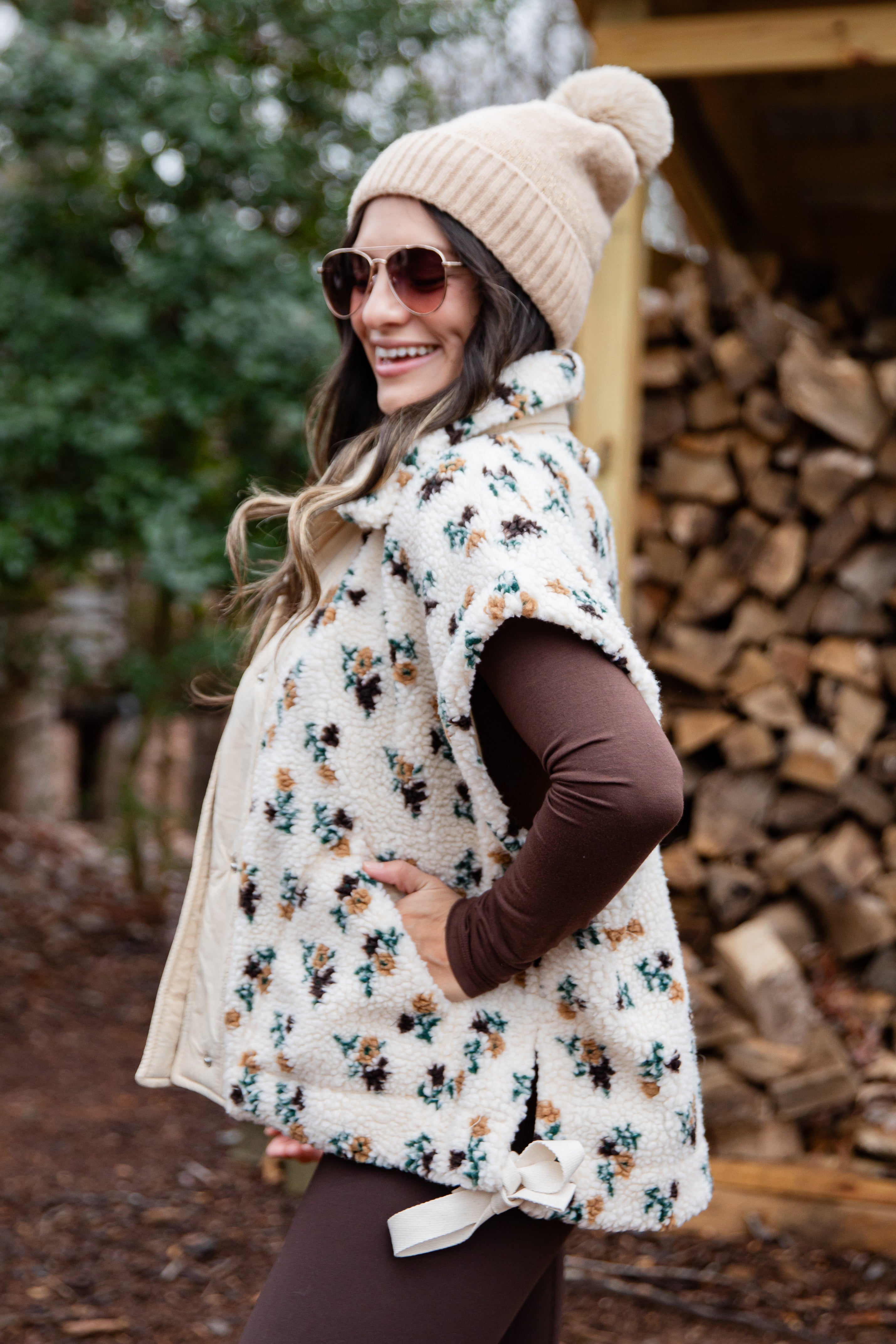 Woman wearing a floral patterned vest and beige knit hat in a forest setting.