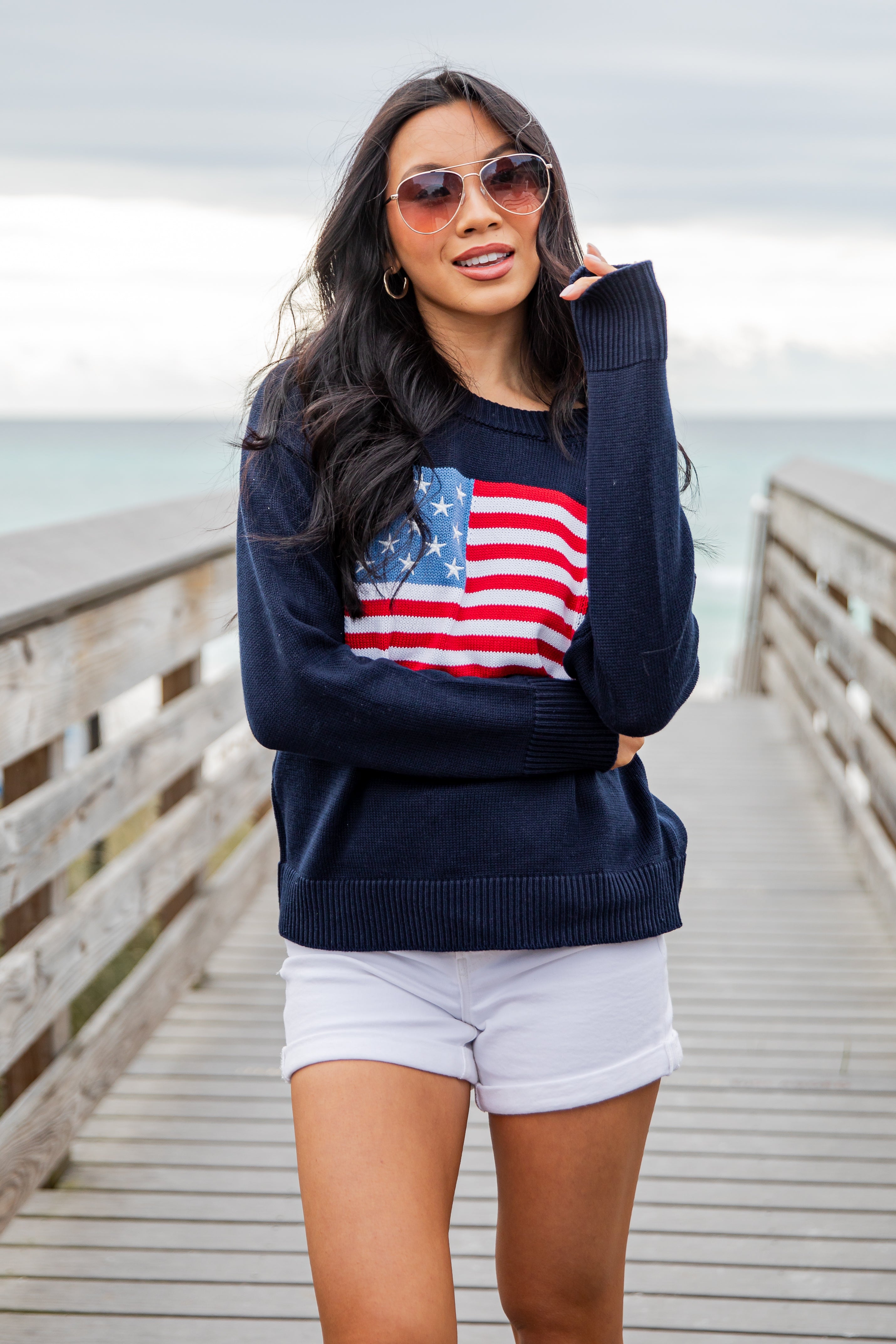 Woman wearing a navy sweater with an American flag design on a wooden pier by the ocean.
