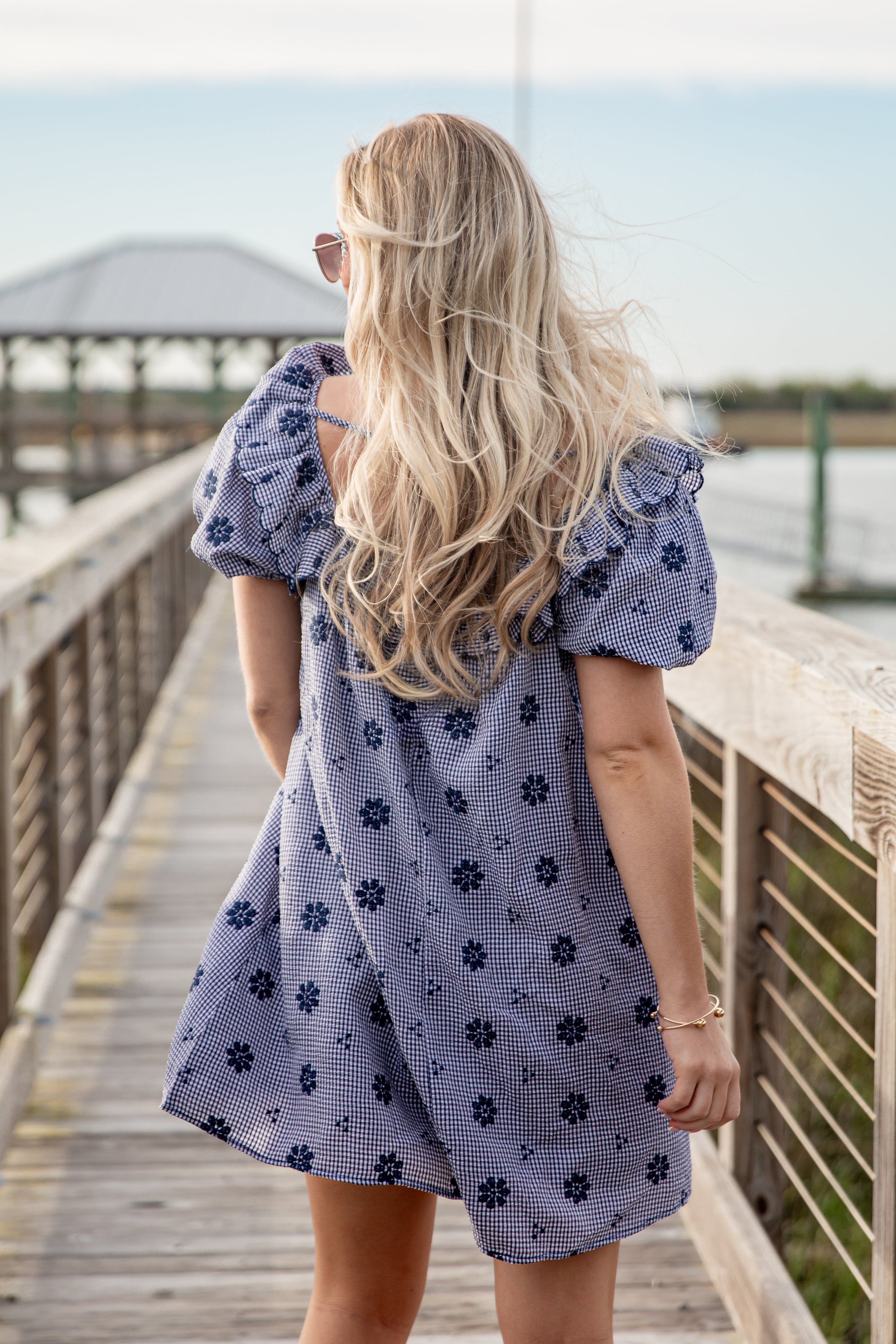 Woman in a blue floral dress standing on a wooden dock by a body of water.