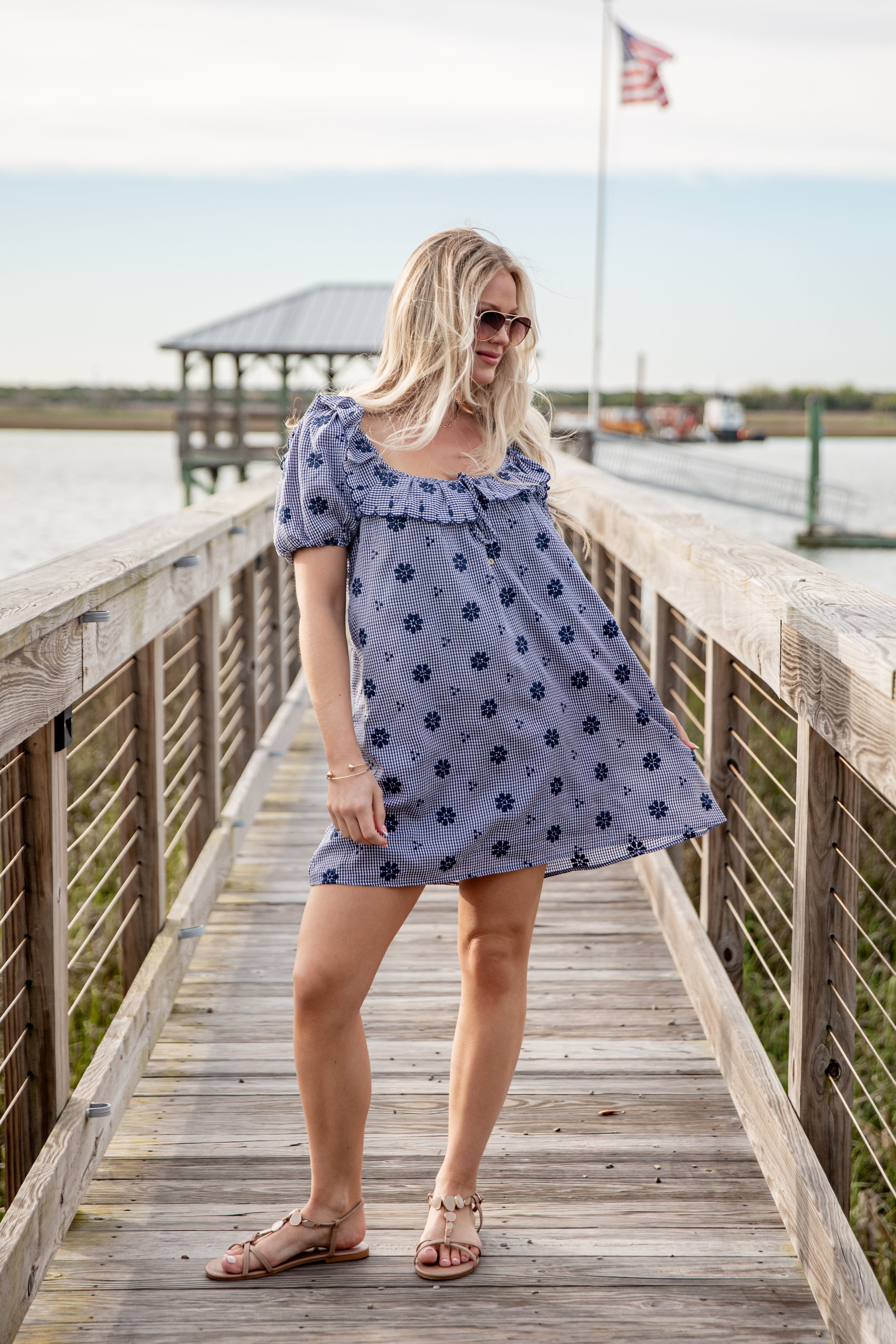 Woman in a blue floral dress standing on a wooden dock by a body of water.