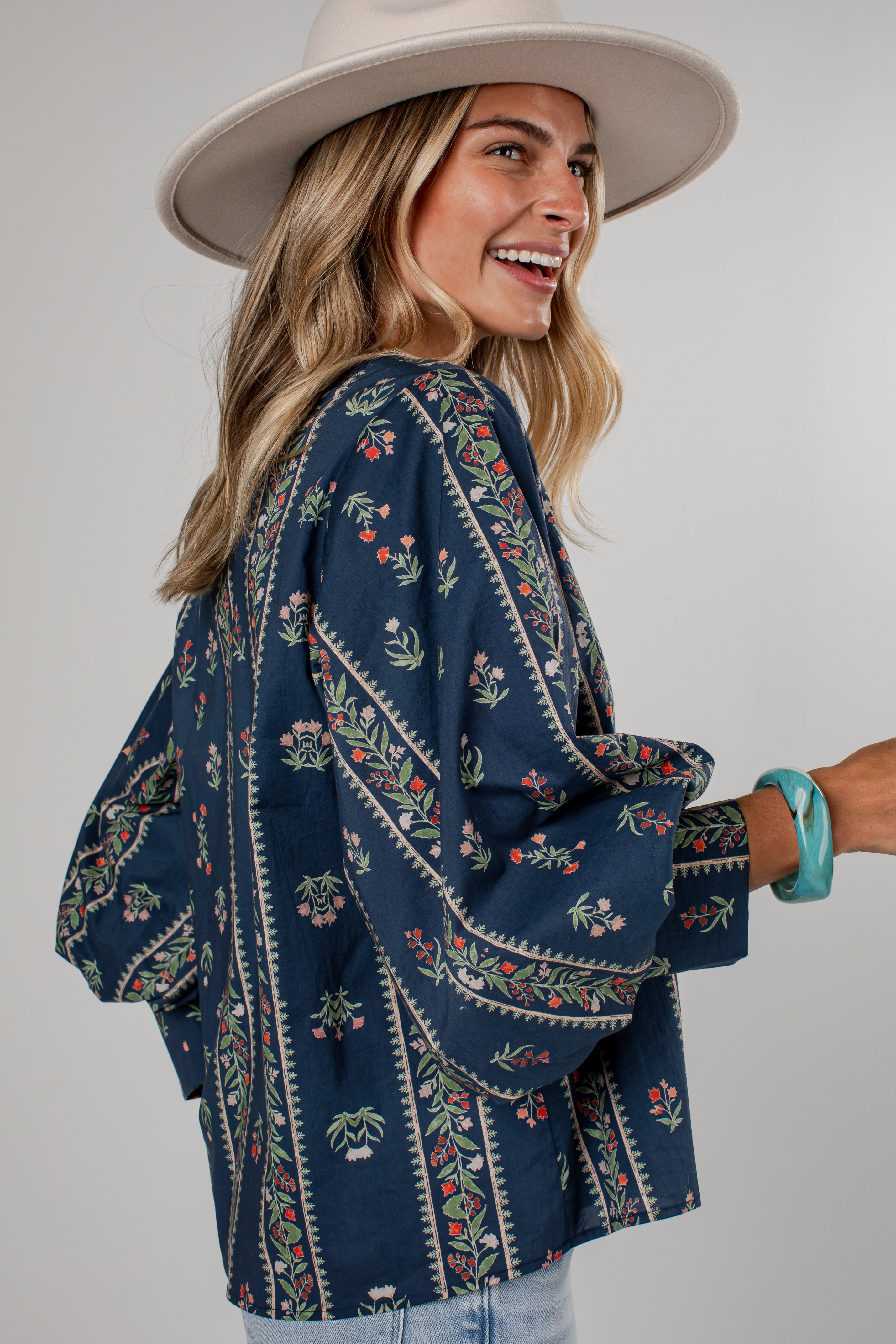 A woman wearing a navy blue bohemian-style embroidered blouse with floral patterns, a wide-brimmed beige hat, and light blue jeans, photographed against a neutral gray studio background.