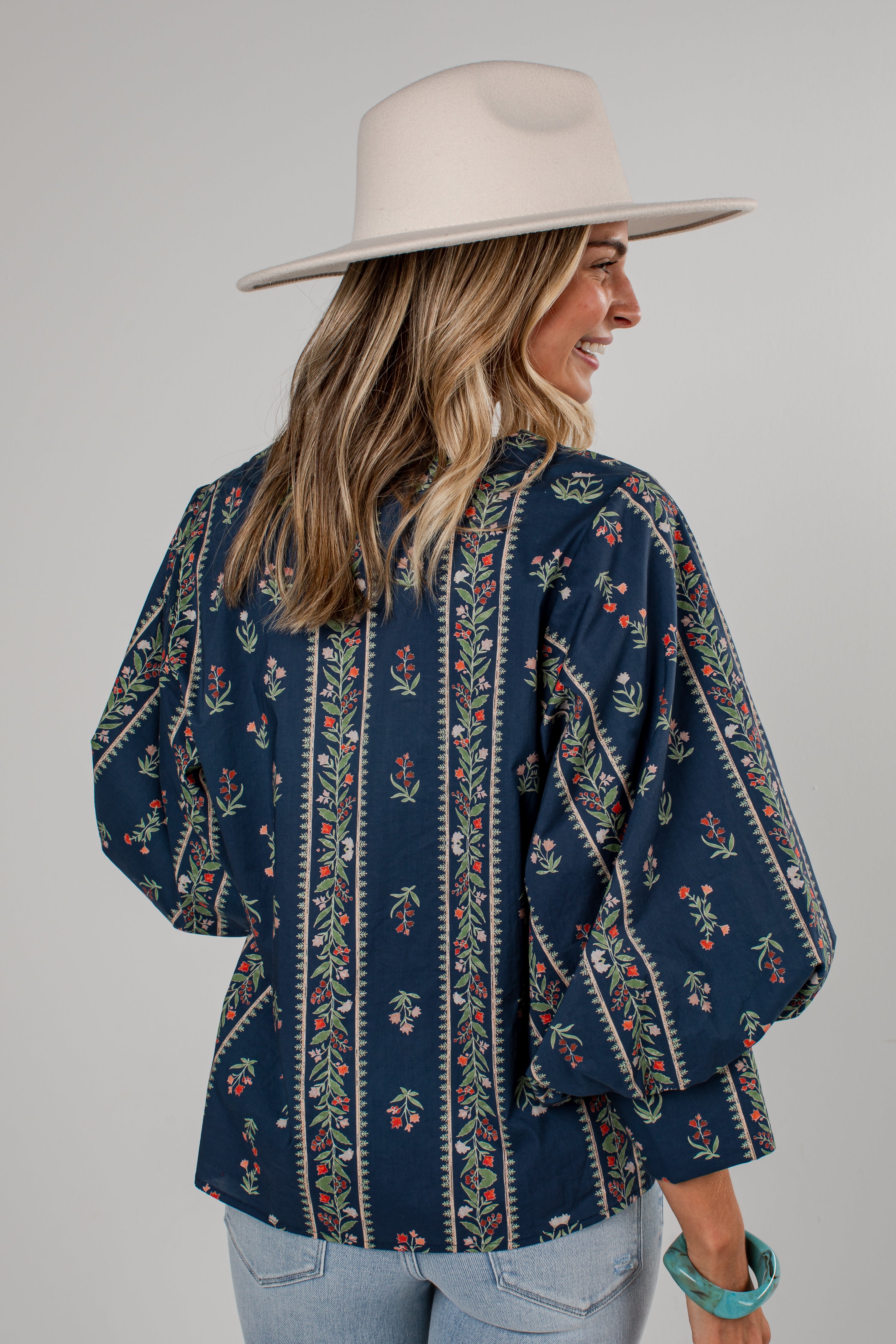 A woman wearing a navy blue bohemian-style blouse with vertical floral and geometric patterns, paired with a wide-brimmed cream hat and light blue jeans, shown from a three-quarters back view against a solid gray background.