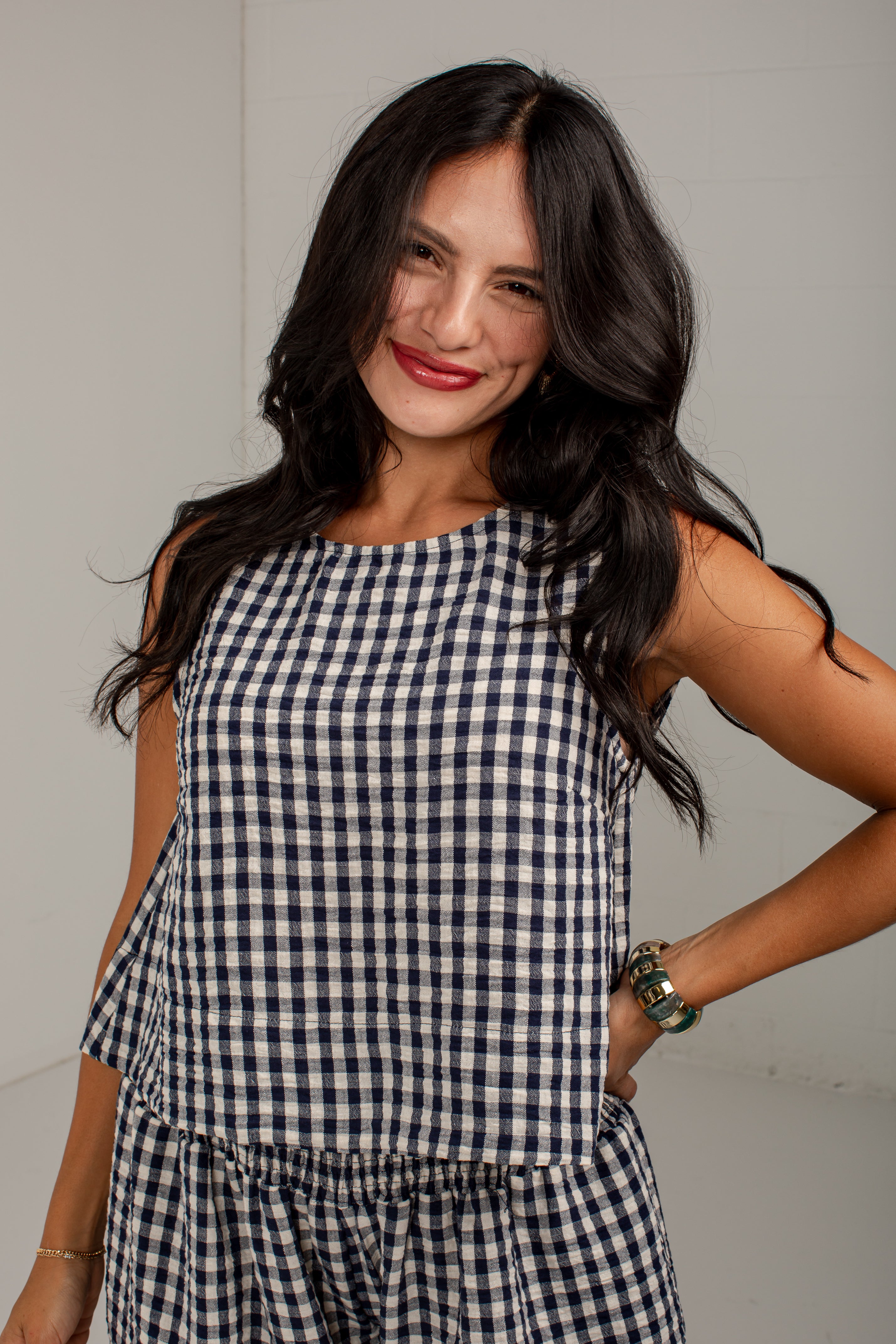 A woman with long dark hair wearing a sleeveless navy and white gingham check top, posing against a plain light gray wall.