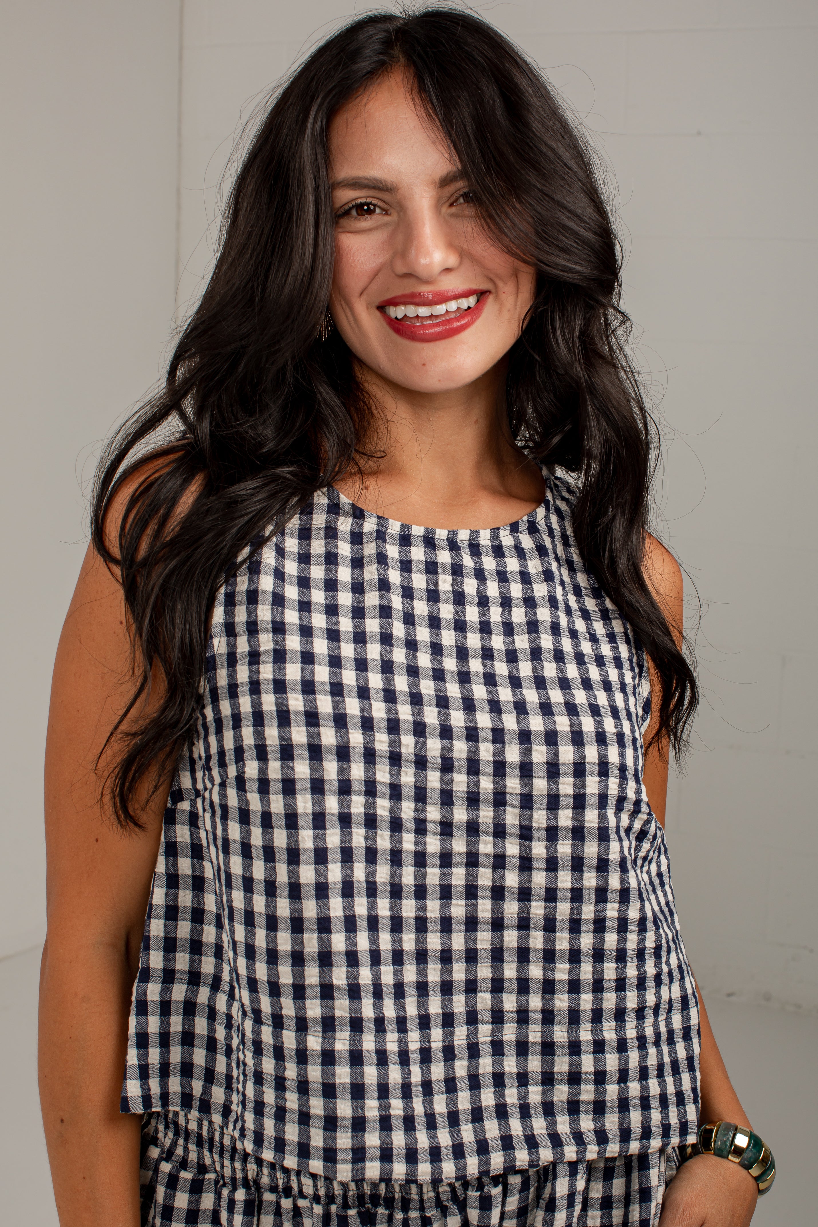 A smiling woman with long dark hair wearing a sleeveless navy and white gingham check top, photographed against a plain light gray wall.