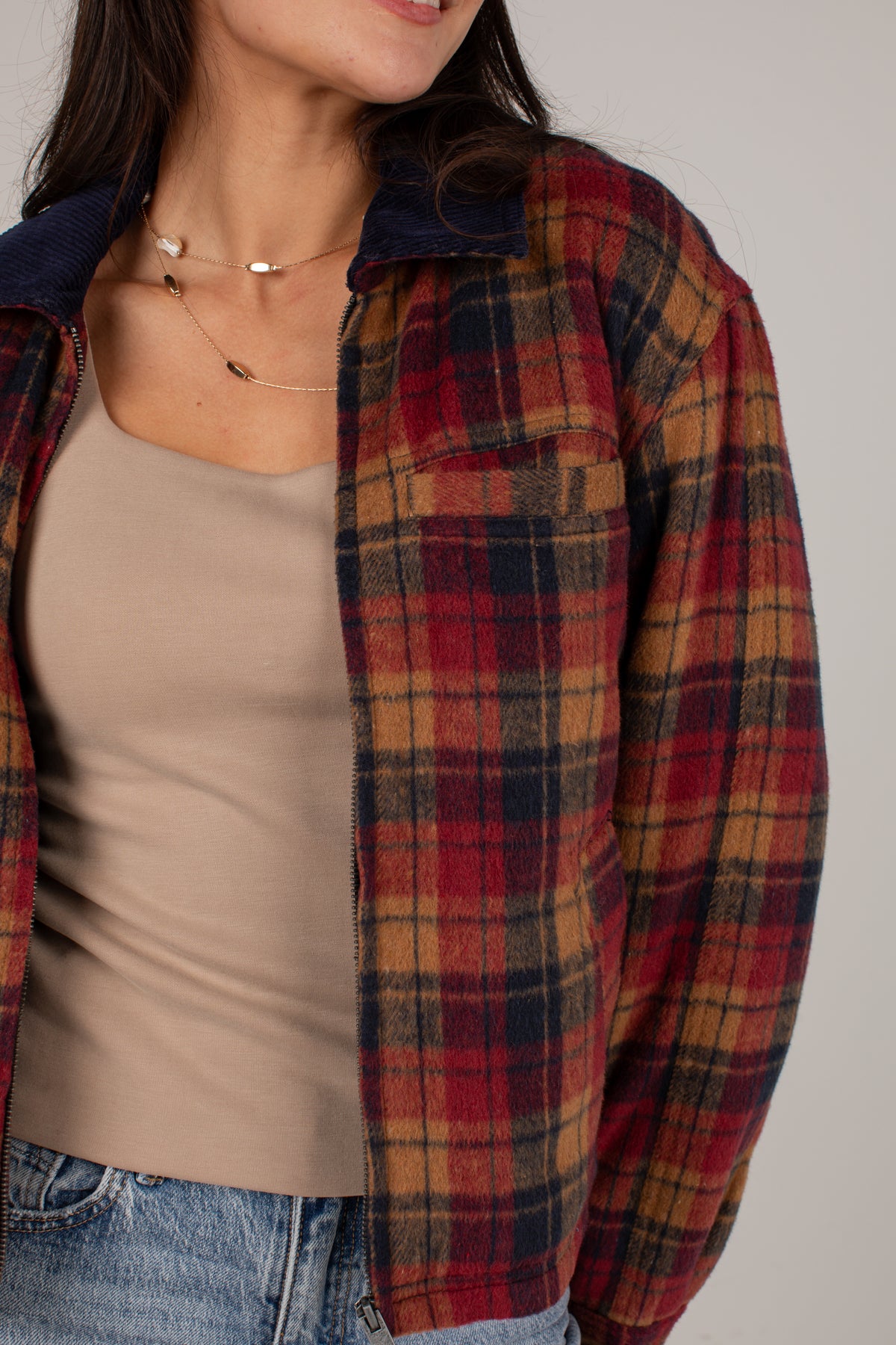 A woman wearing a red, navy, and tan plaid button-up flannel shirt over a beige ribbed tank top and blue jeans, shown from the neck down against a plain light gray background.