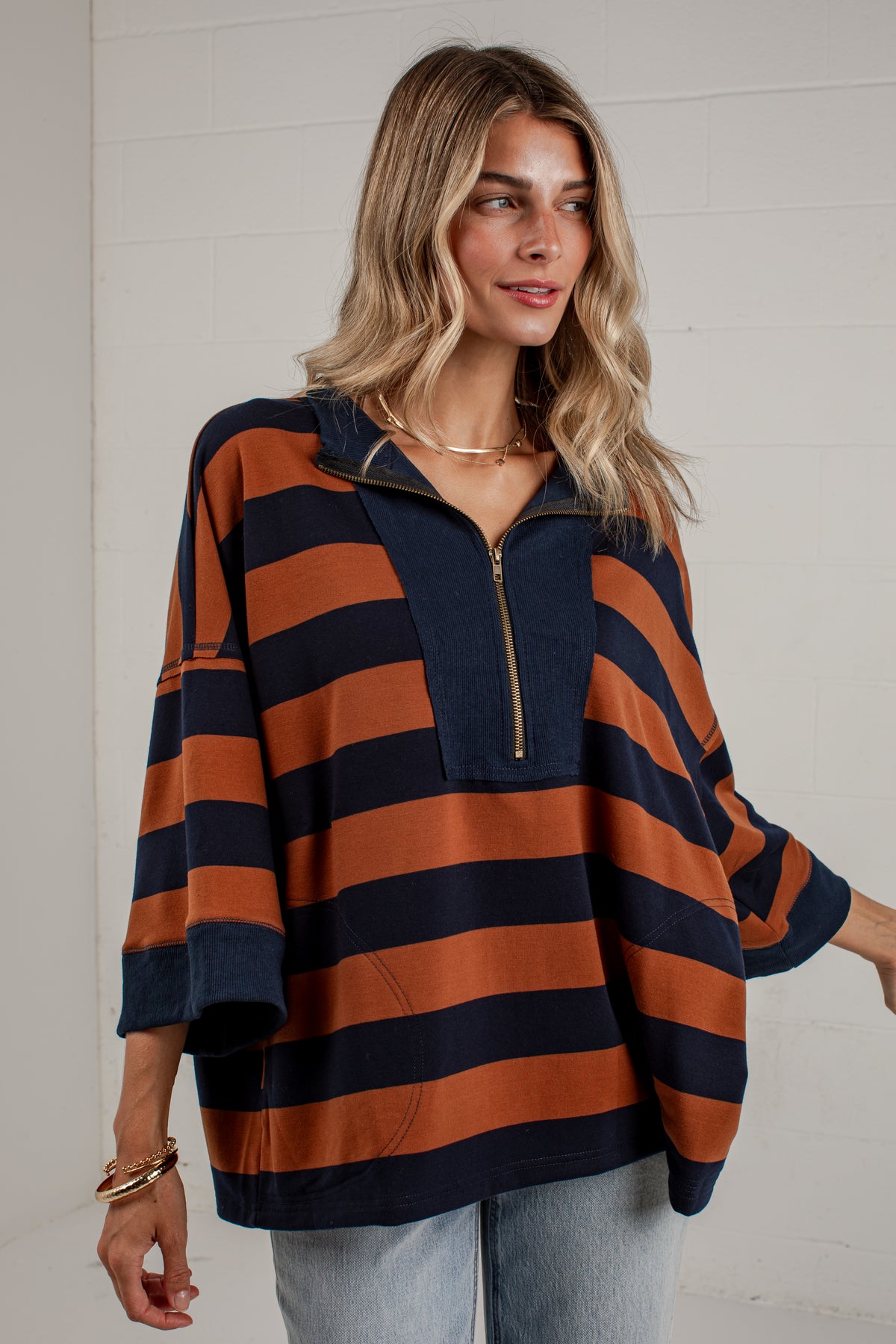 A woman wearing a loose, long-sleeve quarter-zip pullover top with horizontal navy blue and rust-orange stripes, paired with light-wash jeans, standing against a white brick wall.