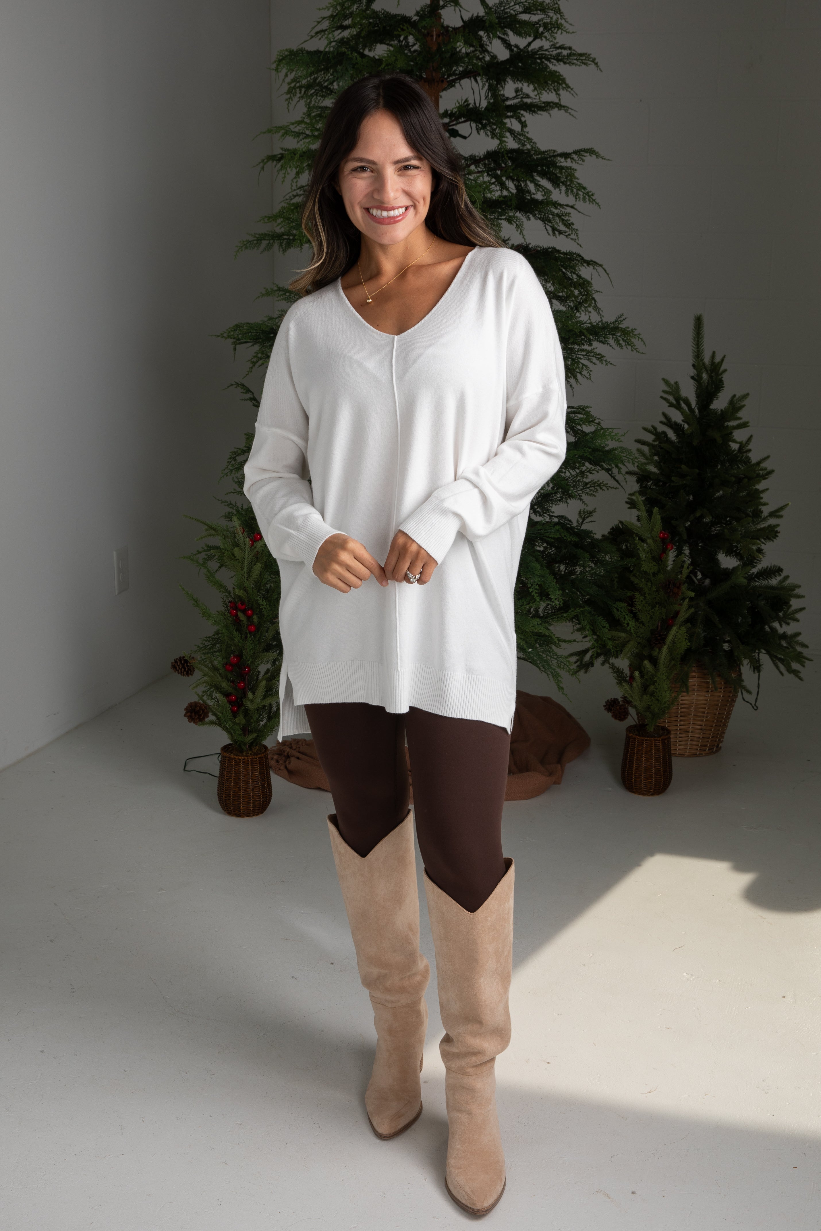 Woman wearing a white long-sleeve top and brown pants with beige boots, standing in front of decorative plants.
