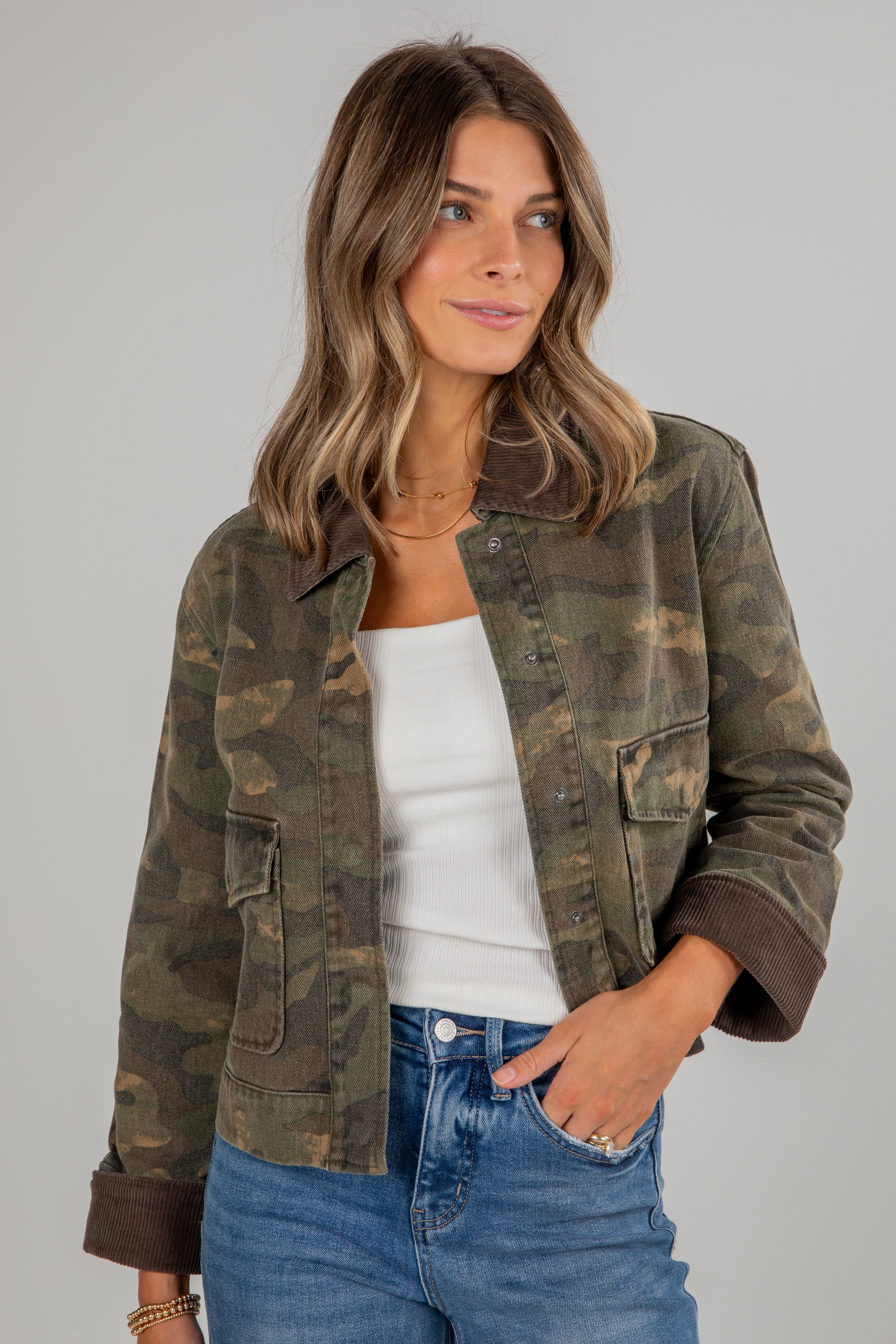 Woman wearing a camouflage jacket over a white top and blue jeans on a gray background