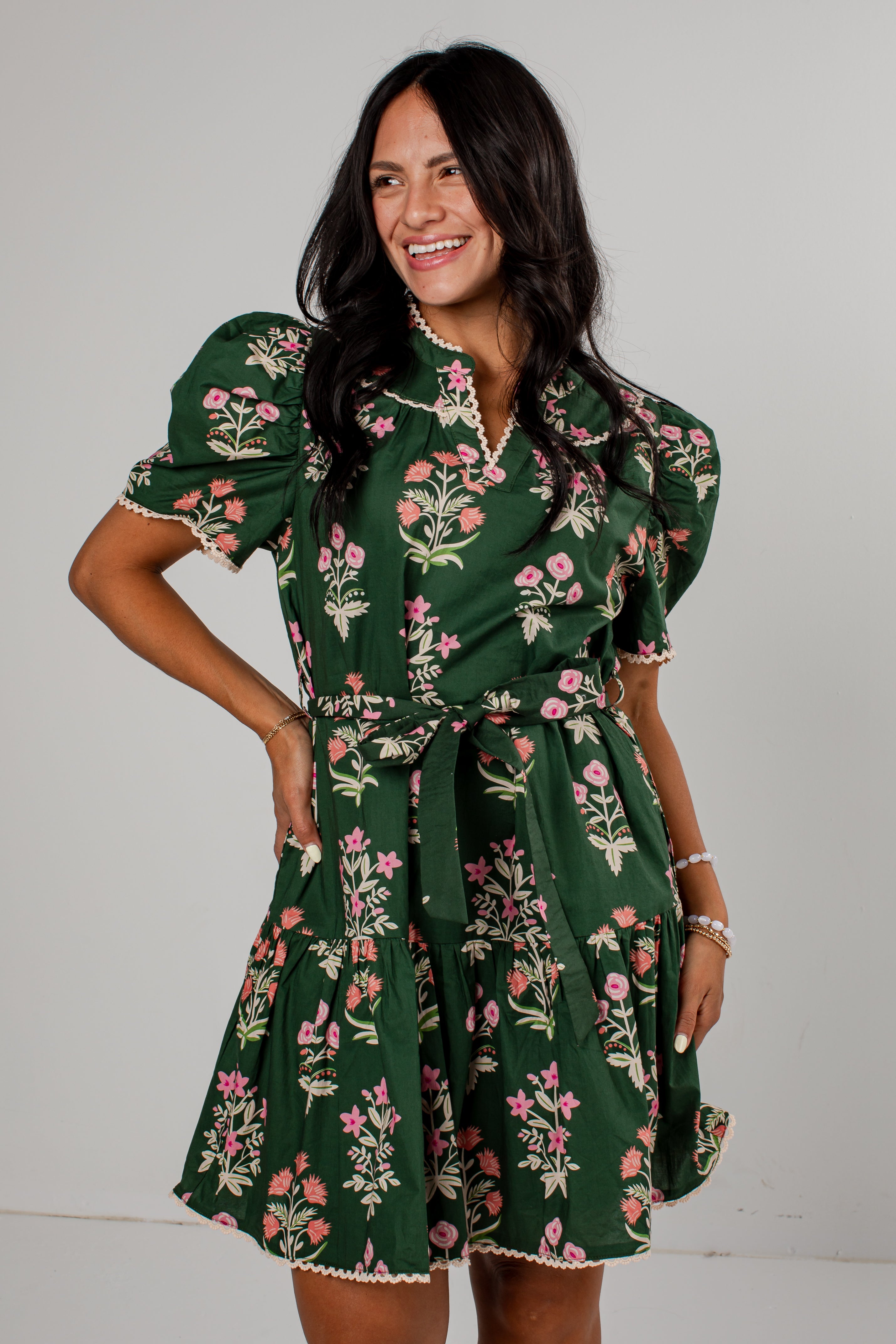A woman modeling a dark green short-sleeve mini dress with a pink and white floral print, featuring puff sleeves, a collar, and a matching fabric tie belt, set against a plain light gray background.