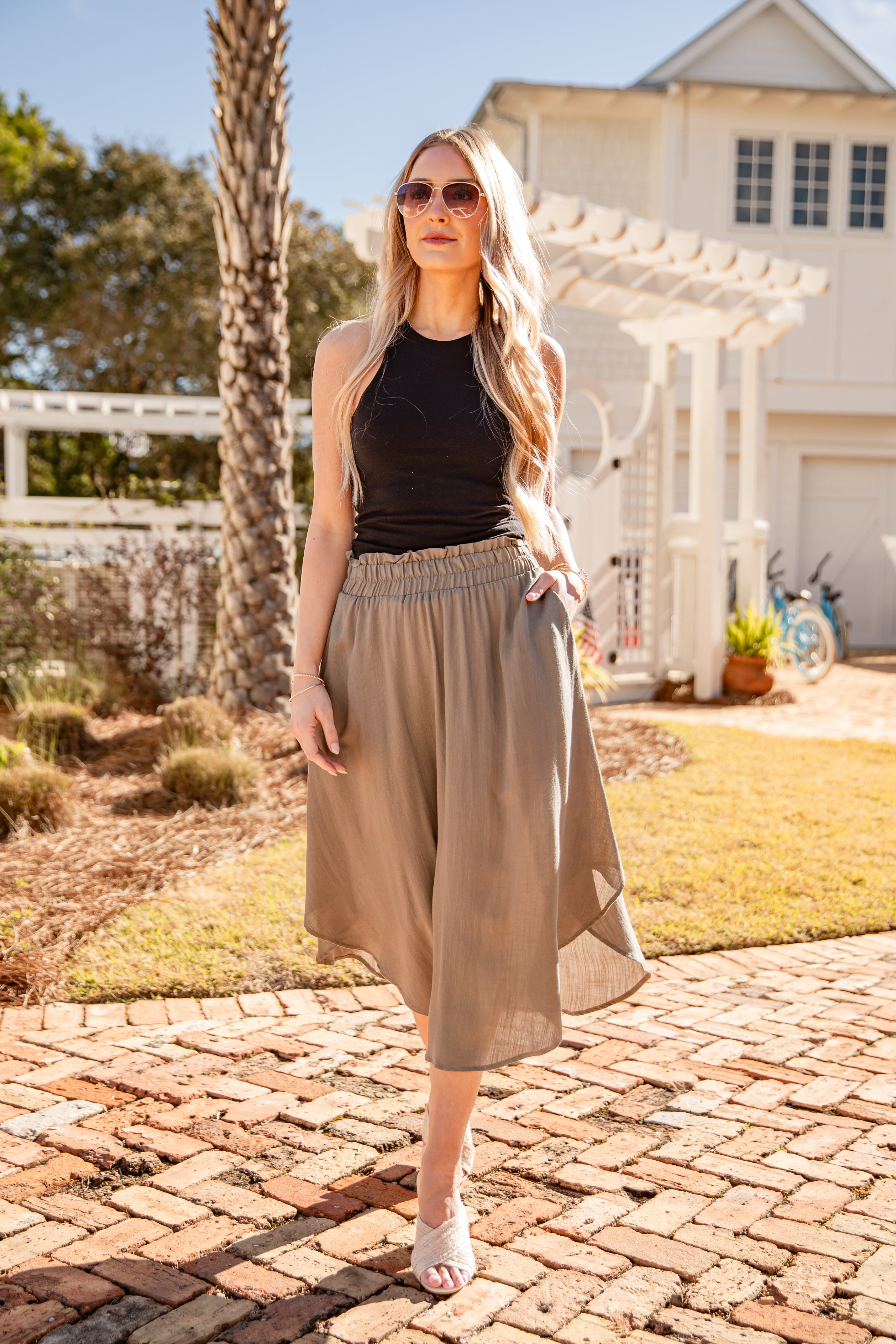 Woman wearing a black top and beige skirt standing on a brick path with a house and trees in the background.