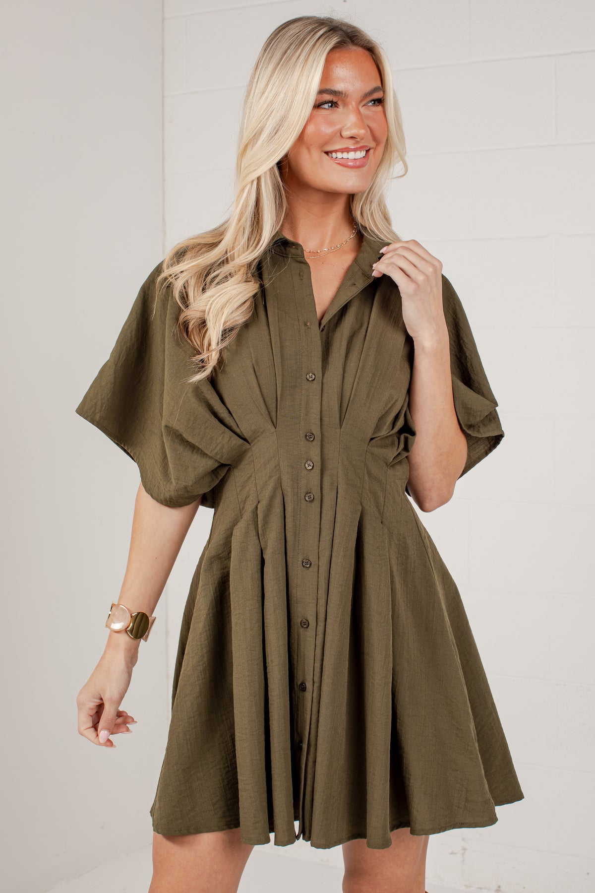 A woman modeling an olive green short-sleeve button-up dress with a flared skirt, standing against a light gray wall.