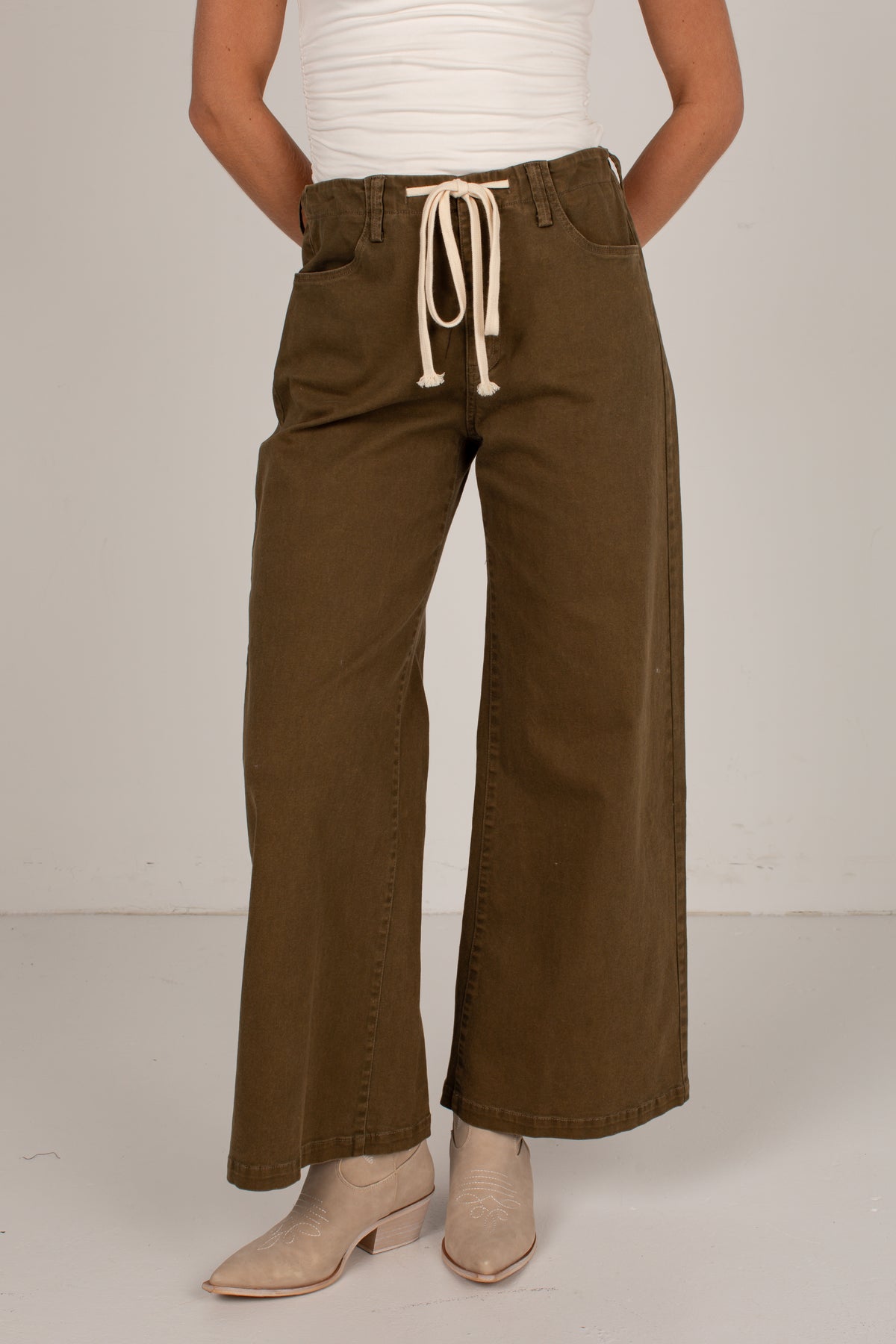 A person wearing wide-leg olive brown pants with a cream drawstring waist, paired with a white ribbed top and beige ankle boots, shown from the waist down against a plain light gray studio background.
