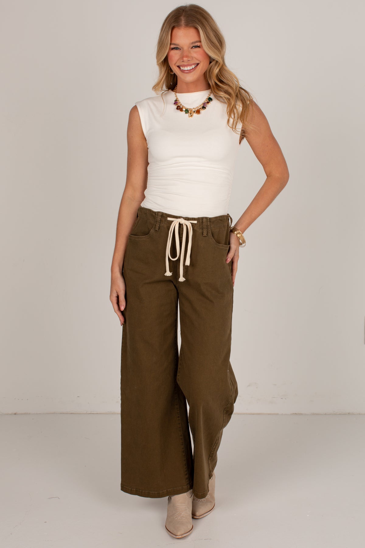 A woman modeling a white sleeveless top and olive green wide-leg drawstring pants against a plain light gray studio background.