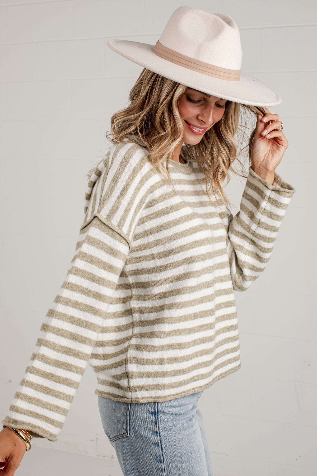 A woman wearing a beige and white striped knit sweater, a cream wide-brim fedora hat, and light blue jeans, posing against a white paneled wall.