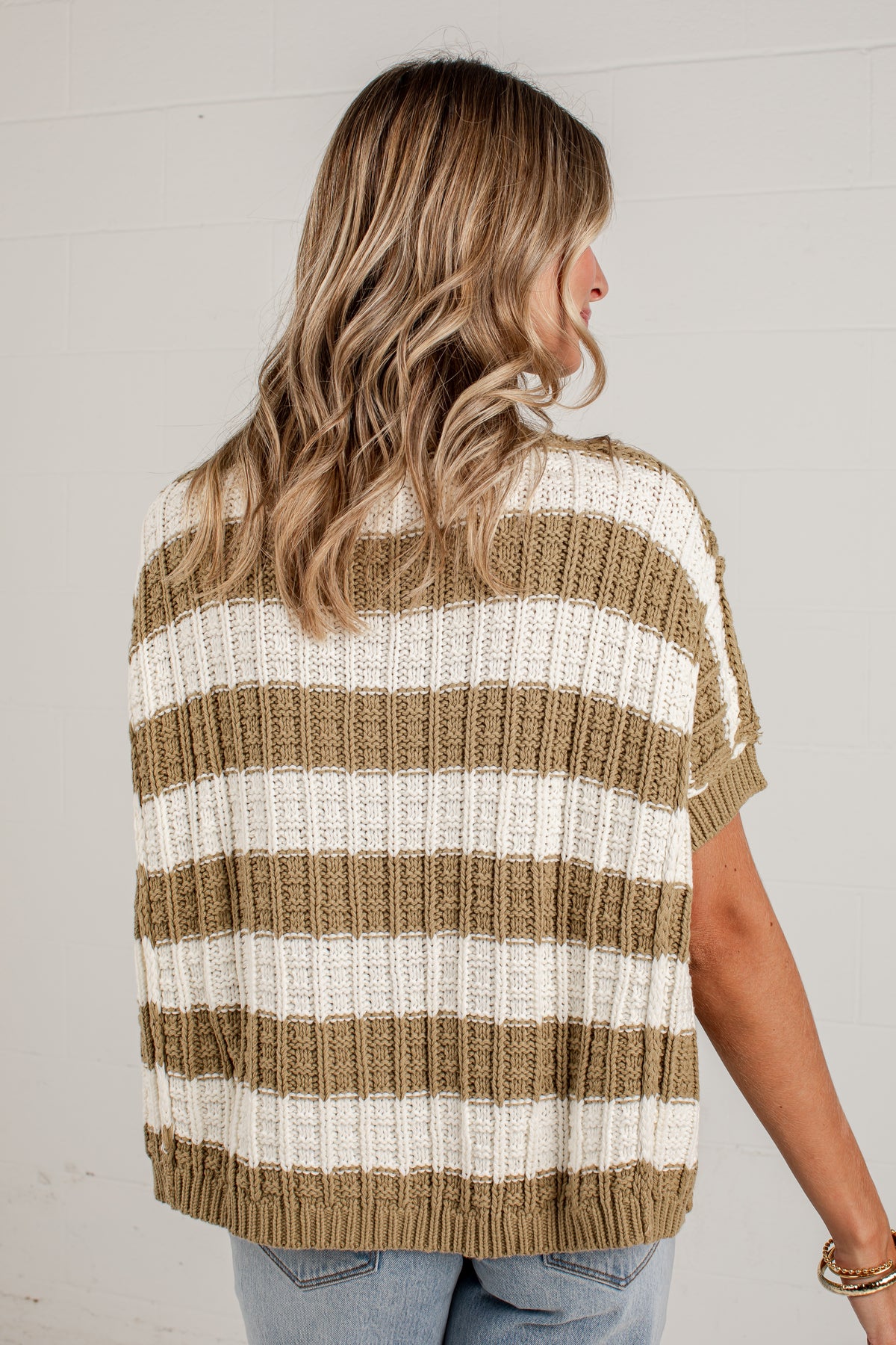 A woman shown from the back wearing a short-sleeve knit sweater with horizontal stripes in tan and off-white, paired with light blue jeans against a white paneled wall.