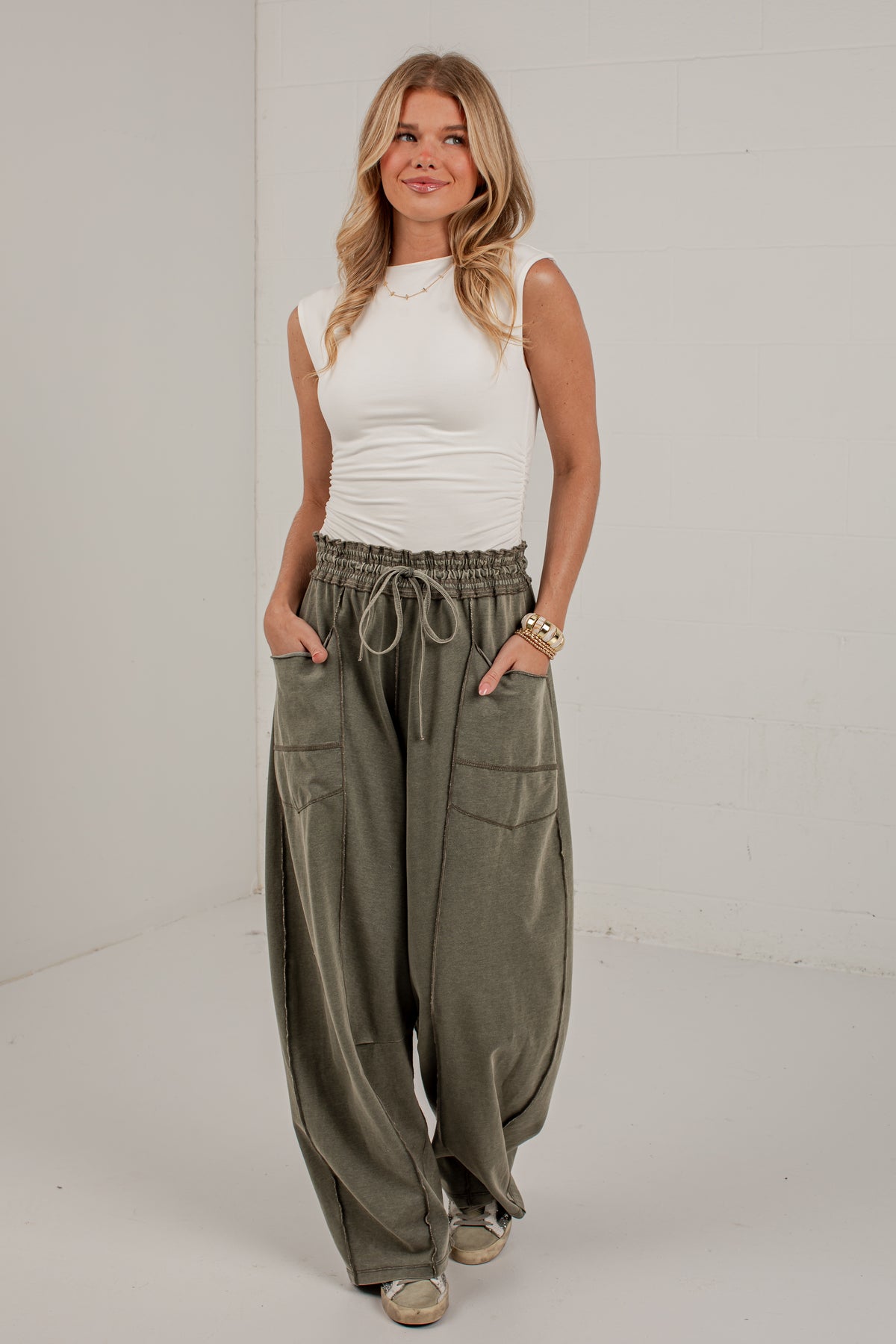 A woman modeling a pair of loose, wide-leg olive green cargo pants with a drawstring waist, paired with a white sleeveless top, in a bright studio setting.
