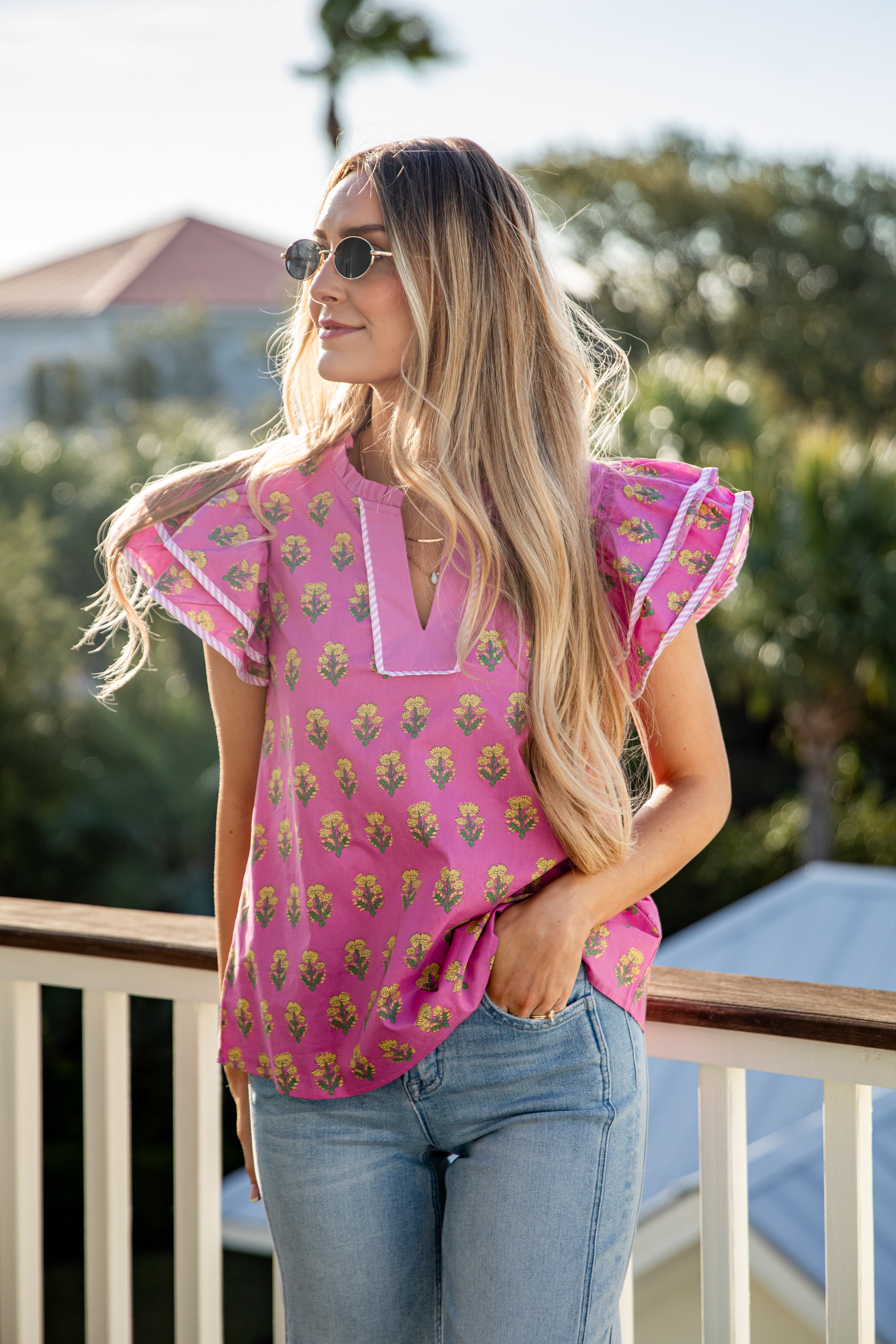 Woman wearing a pink patterned top and blue jeans on a balcony with greenery in the background