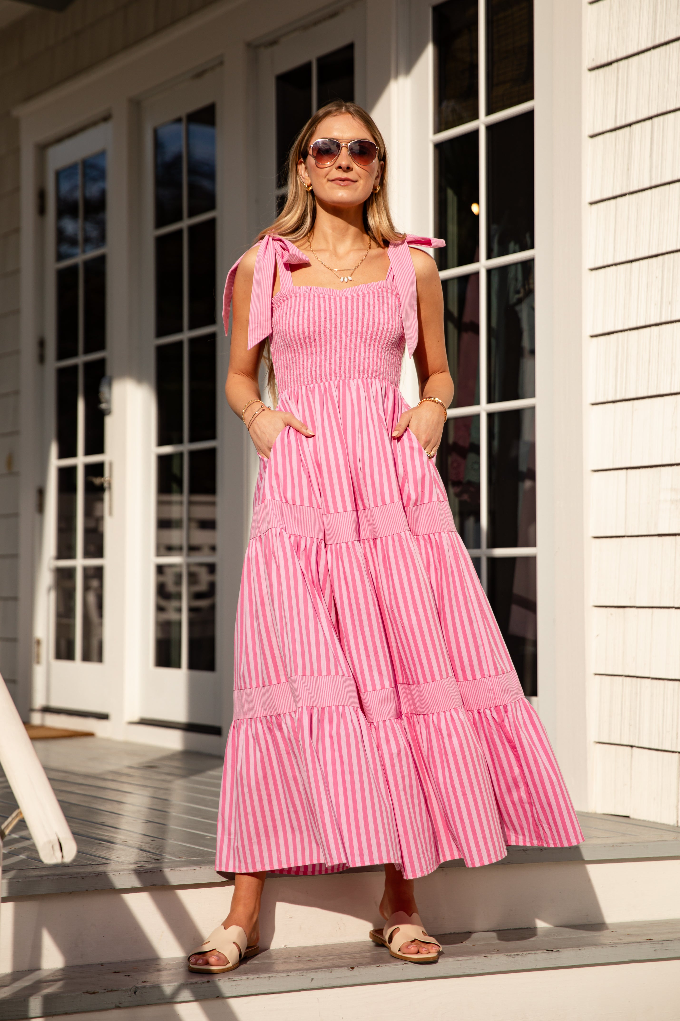 Woman wearing a pink striped dress with bow straps standing on a porch.