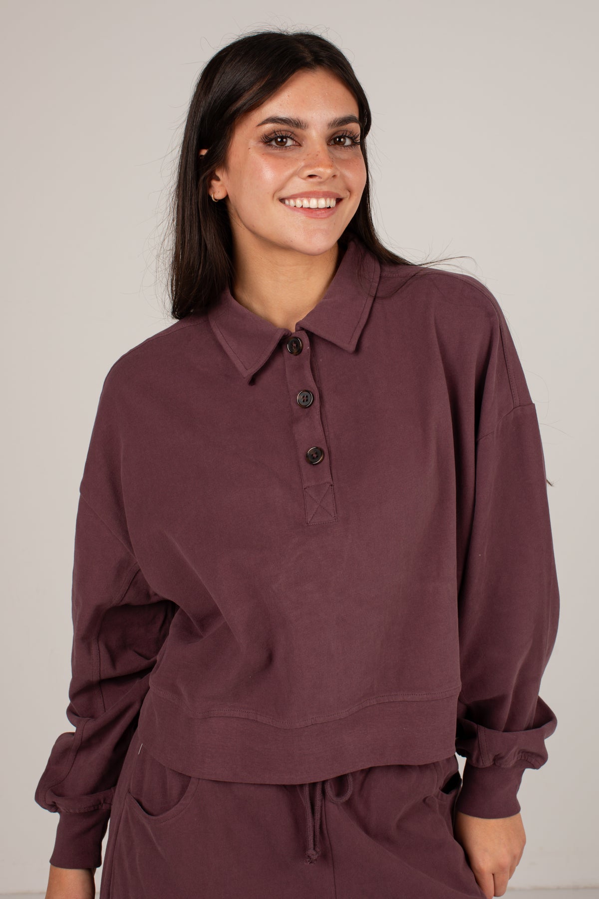 A woman wearing a dark plum-colored long-sleeve polo shirt with a buttoned collar, paired with matching pants, photographed against a neutral light gray background.