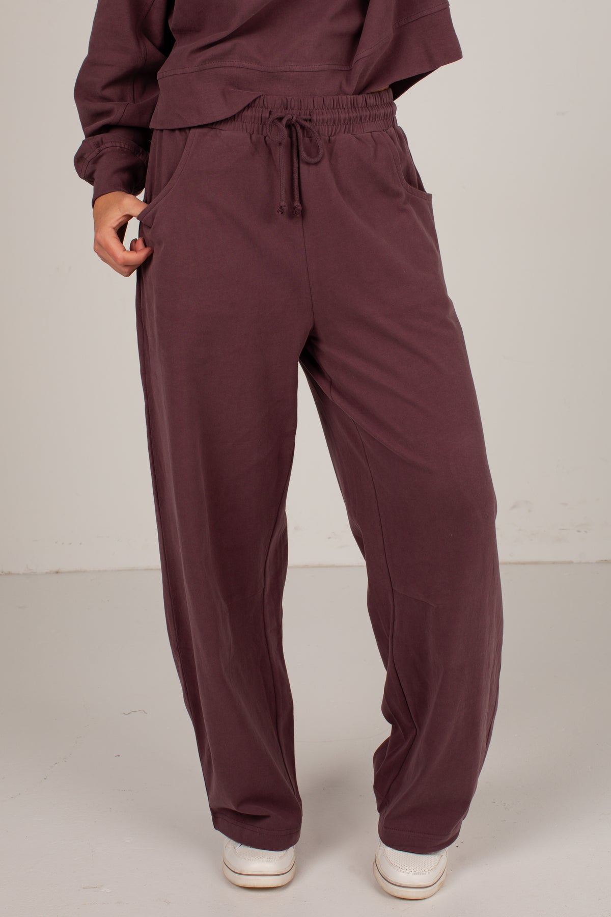 A person wearing loose, wide-leg drawstring sweatpants in a deep plum-burgundy color, paired with white sneakers against a plain light gray studio background.