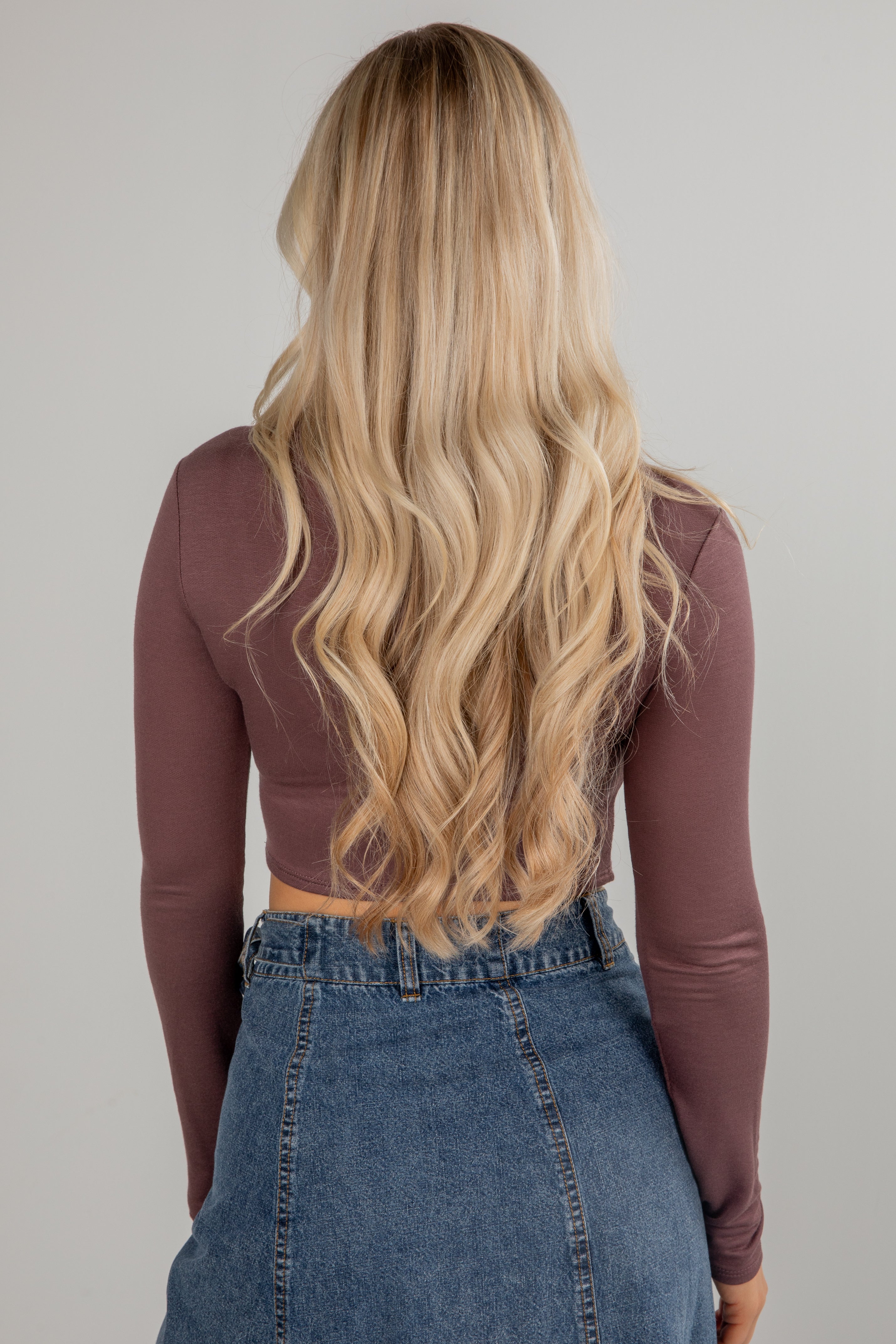Person with long blonde hair wearing a brown long-sleeve top and blue jeans on a plain background