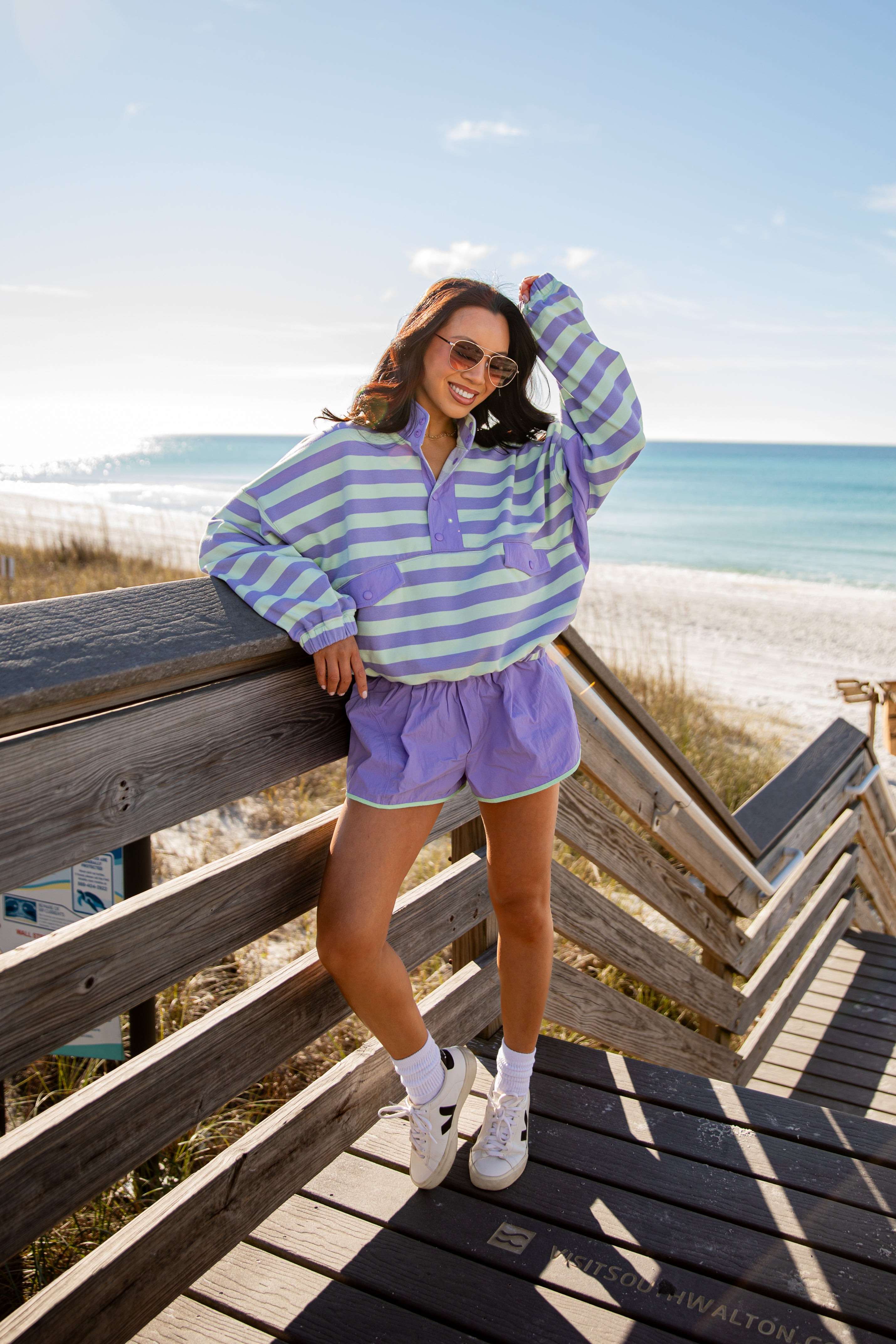 Woman in a striped sweater and shorts standing on a wooden deck by the beach.