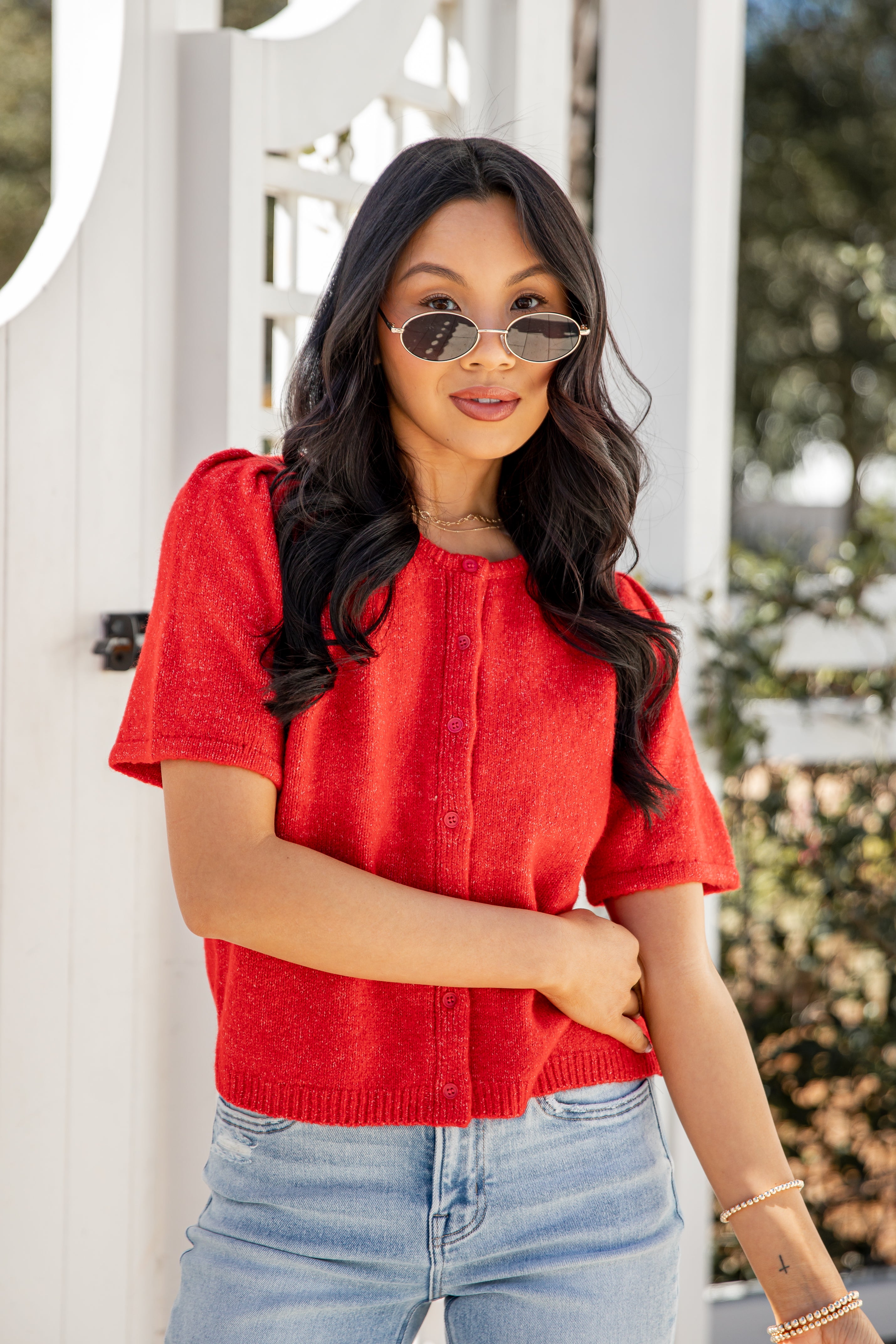 Woman wearing a red top and sunglasses standing in front of a white gate.