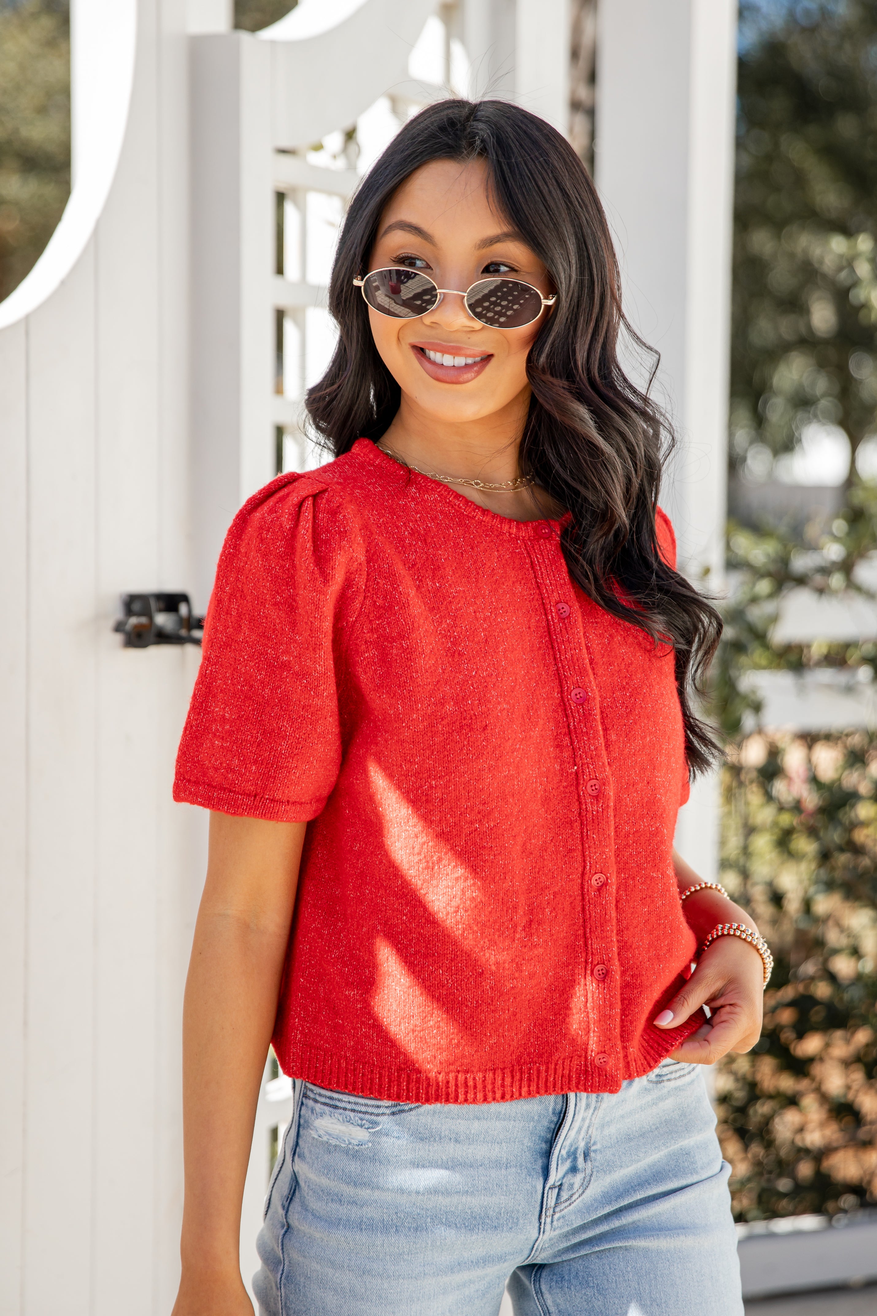 Woman wearing a red top and sunglasses, standing in front of a white door.