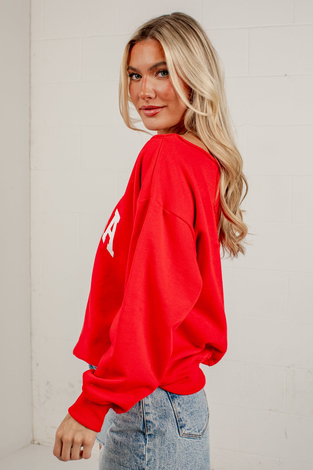A blonde woman wearing a bright red oversized crewneck sweatshirt with a white graphic letter on the sleeve, paired with light-wash blue jeans, standing against a white brick wall.