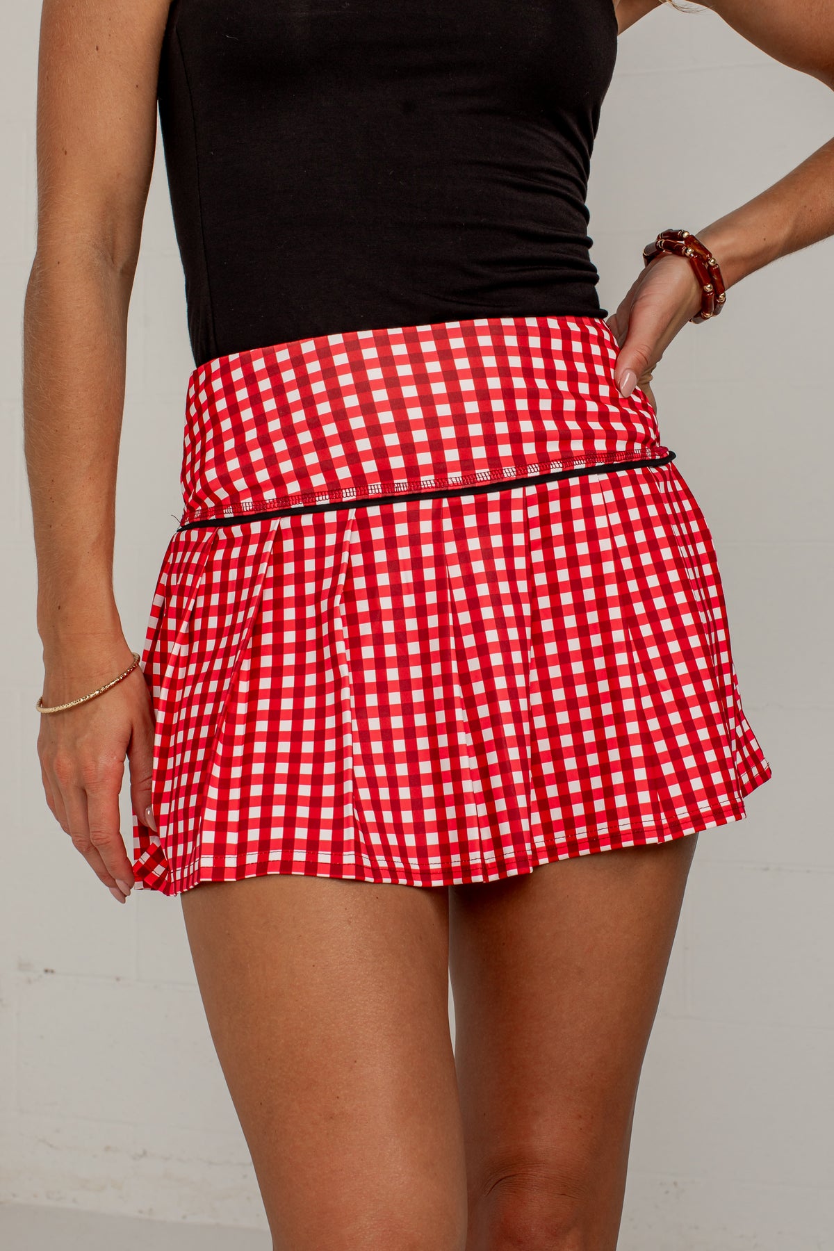 A close-up of a model wearing a red and white gingham pleated tennis skirt with a black waistband trim, paired with a black sleeveless top.